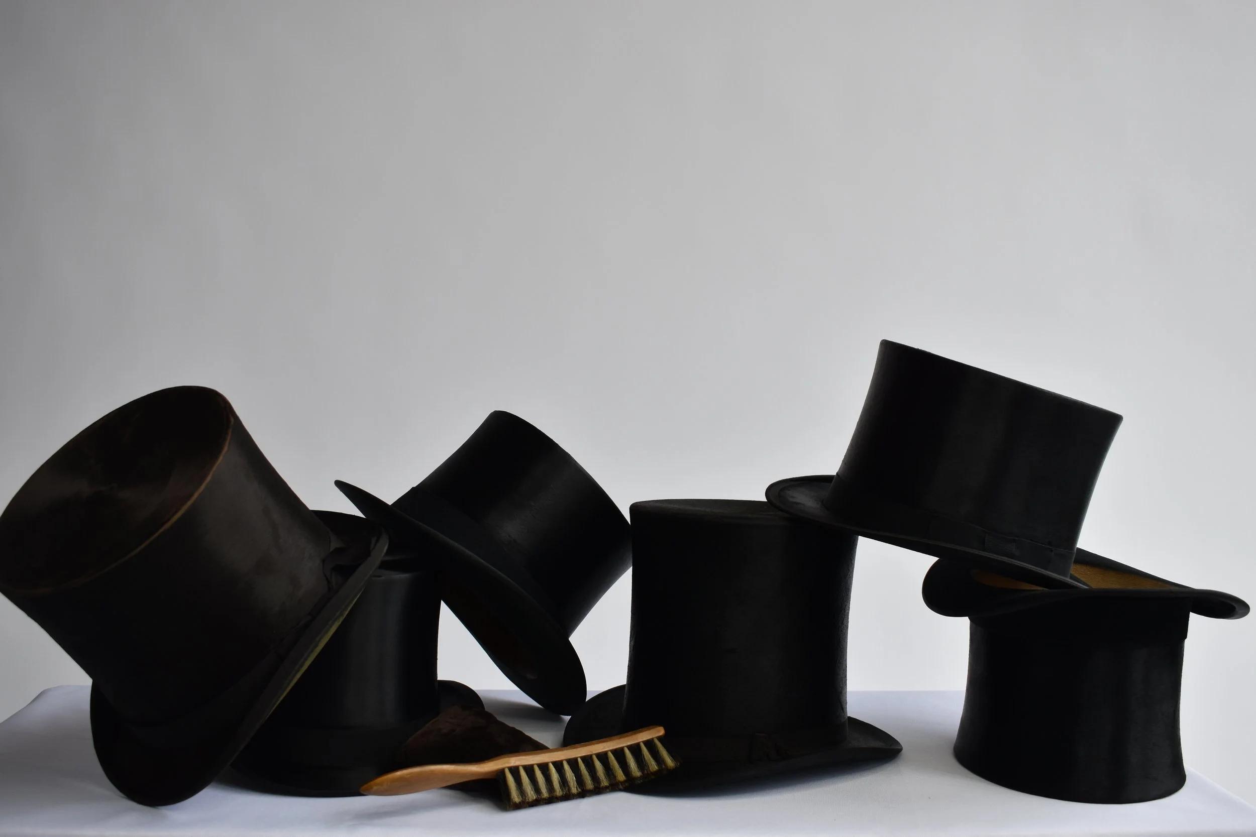 A collection of black top hats stacked and scattered with a wooden hairbrush and comb lying in front on a white surface against a grey background.
