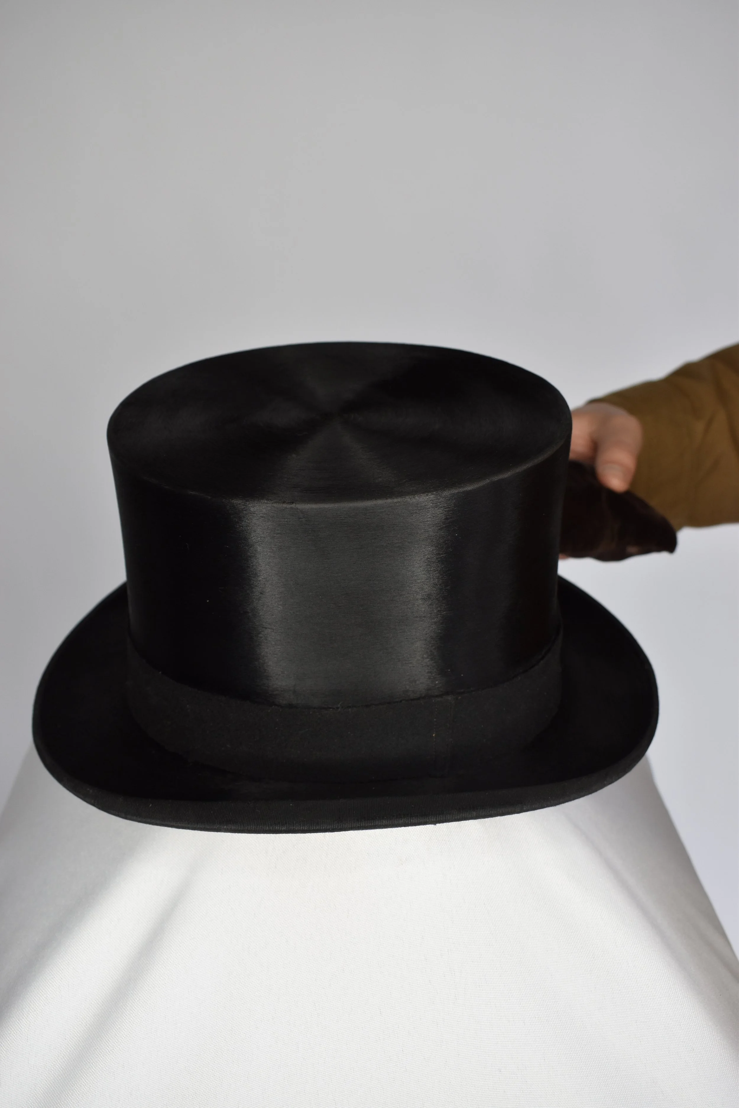 Black top hat with a flat crown and wide brim, placed on a white surface, with the hand of a person wearing a brown jacket visible in the background.