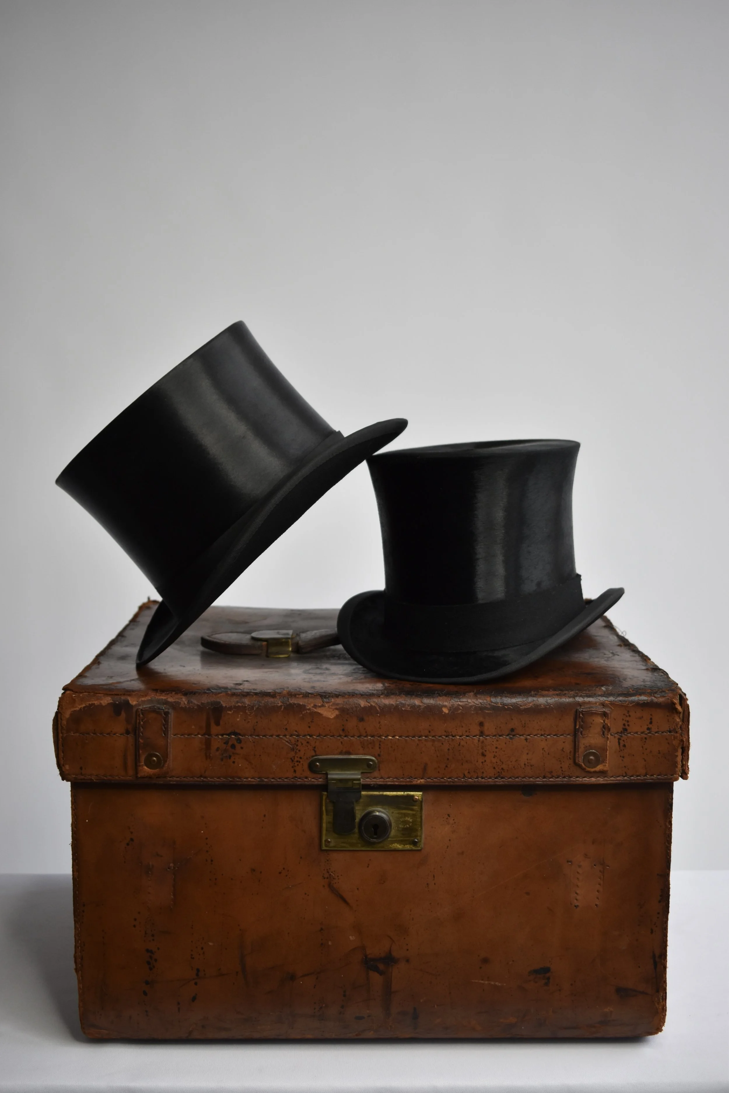 Two black top hats resting on a vintage brown leather suitcase with a brass lock, set against a plain gray background.