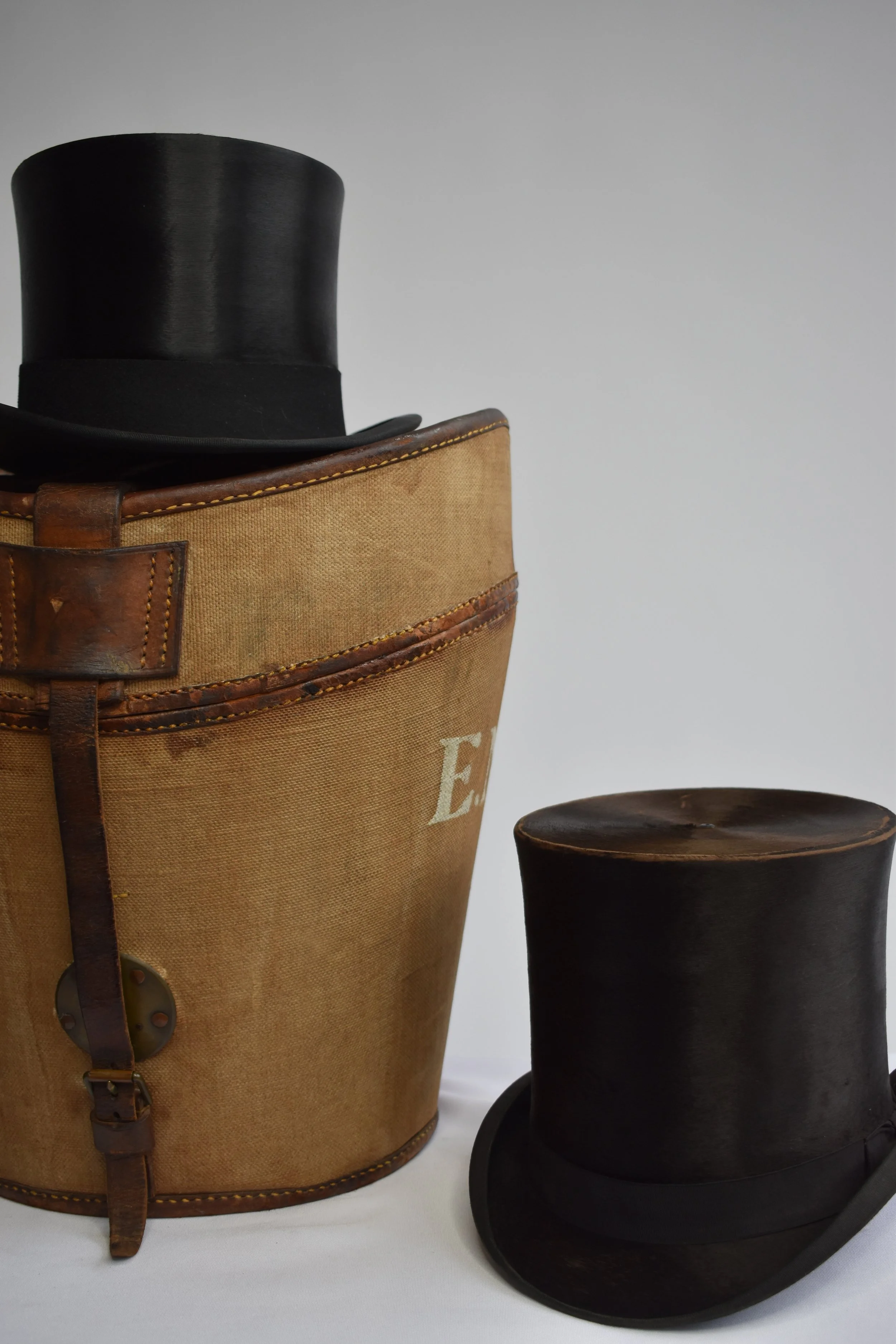 A vintage brown leather and canvas schoolbag with a black top hat inside, and a black colonial hat with a ribbon on a black stand.