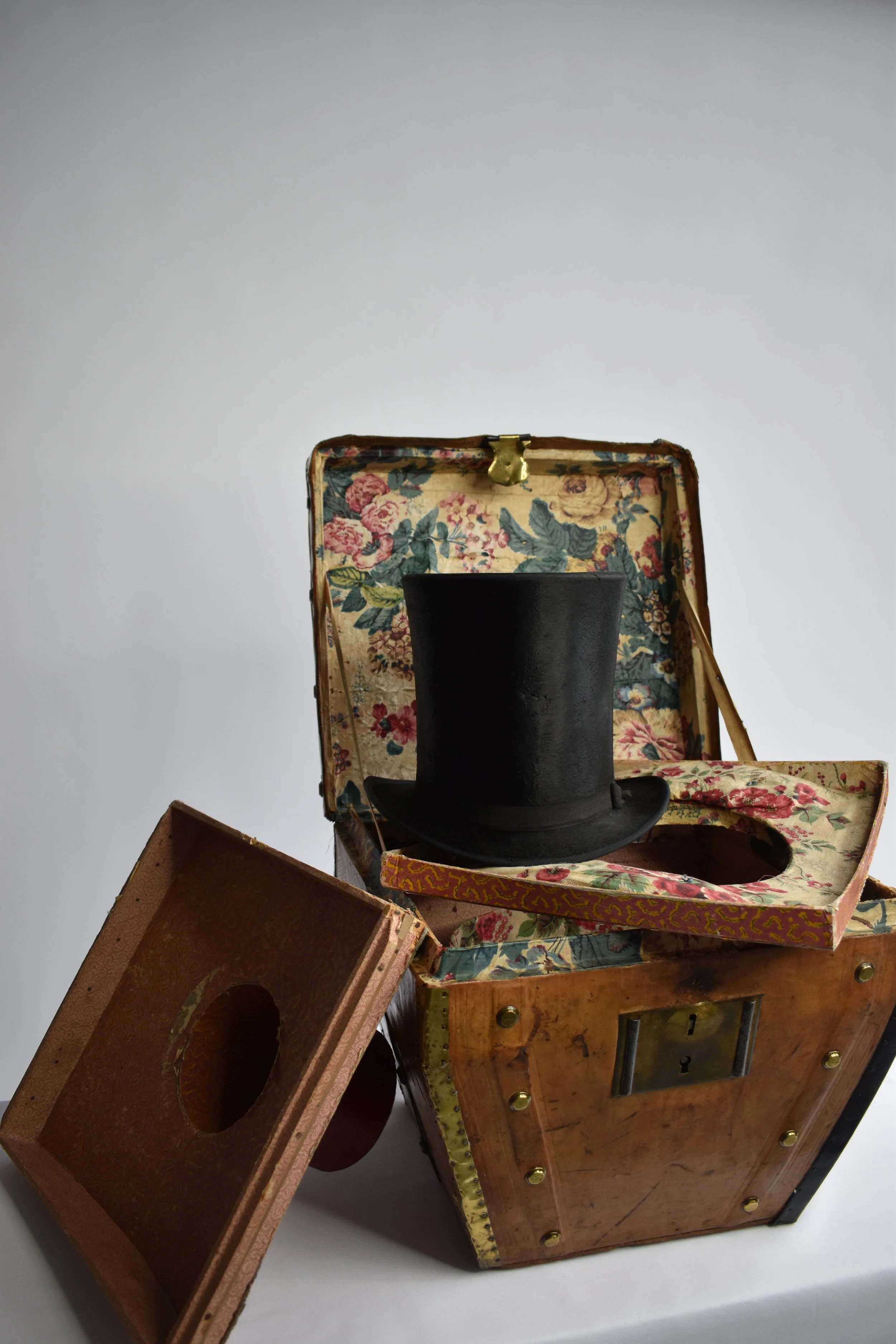 A vintage-style jewelry box with floral fabric lining and a brass clasp, stacked on top of a large wooden box with gold accents and metal hardware. A black top hat is placed inside the jewelry box.