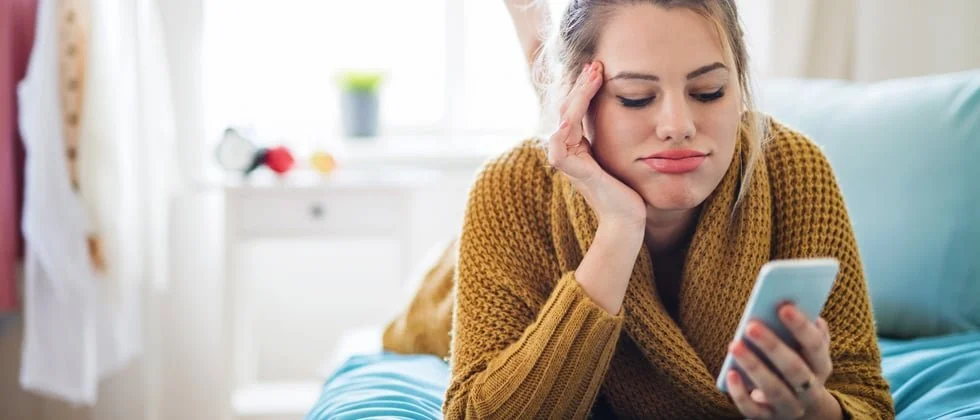 A woman lying on a bed looking at her smartphone with a bored or frustrated expression.