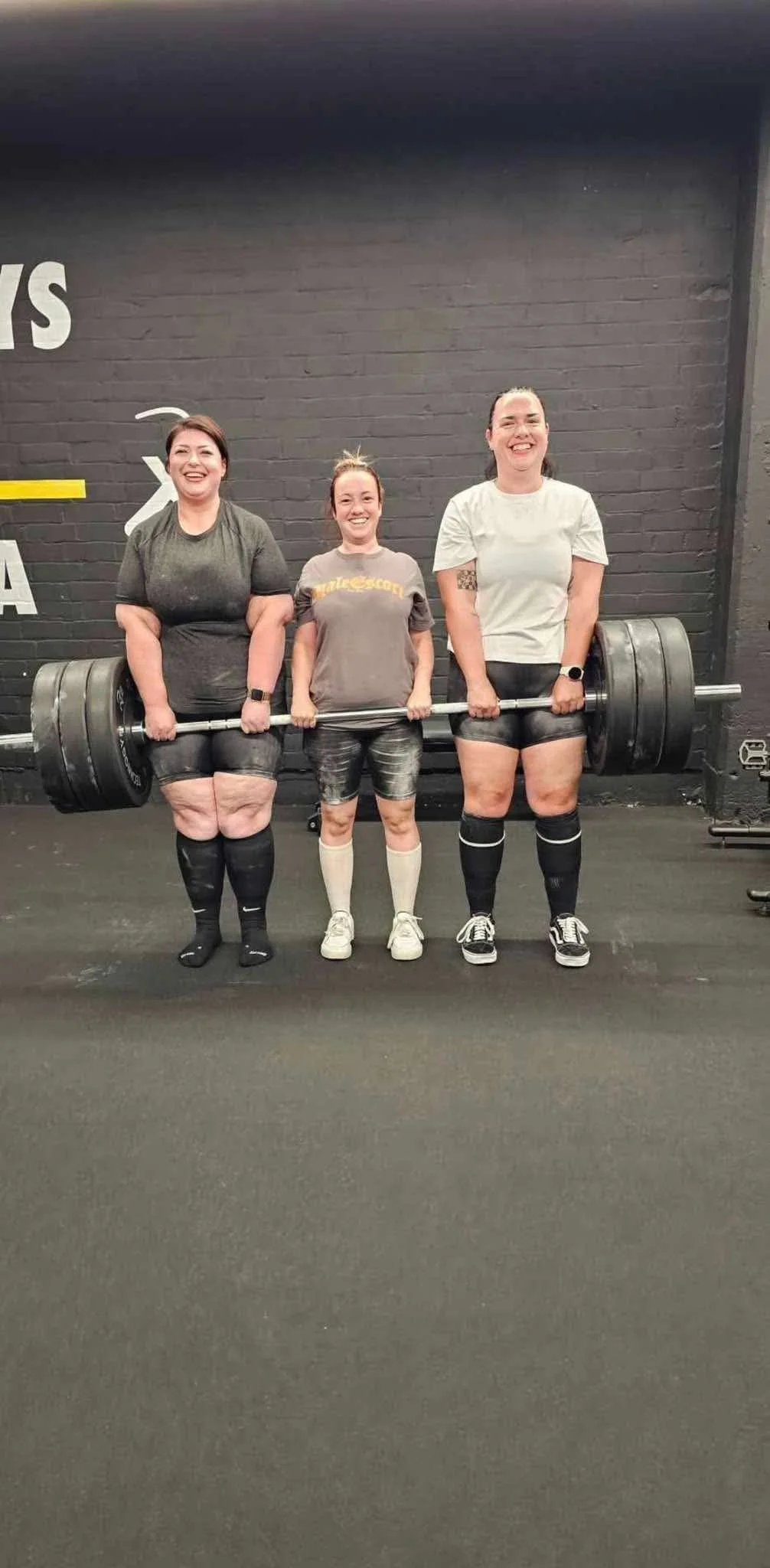 Three women standing in a gym, with two of them holding a heavily loaded barbell overhead, smiling at the camera.