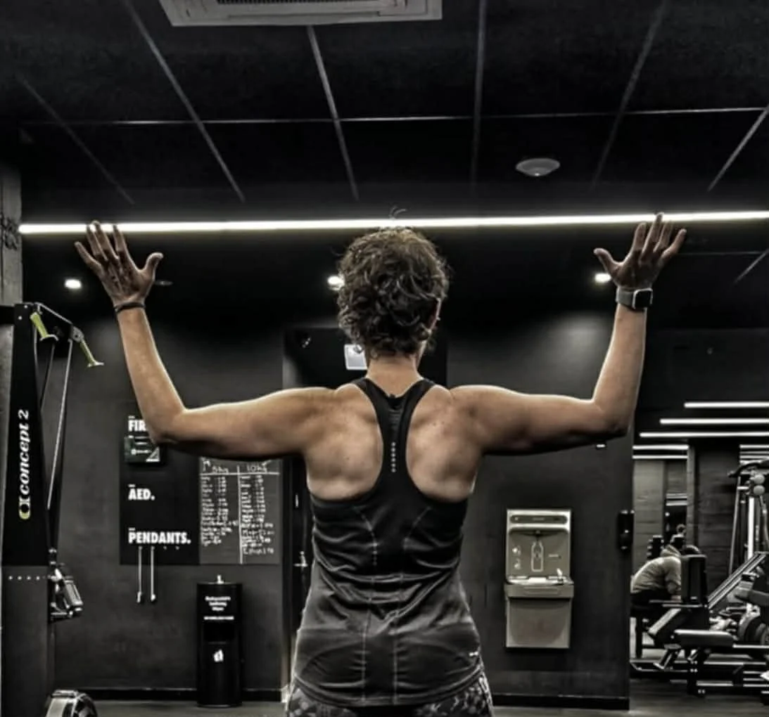 A woman in athletic clothing performing a back workout in a gym, facing away with arms raised.