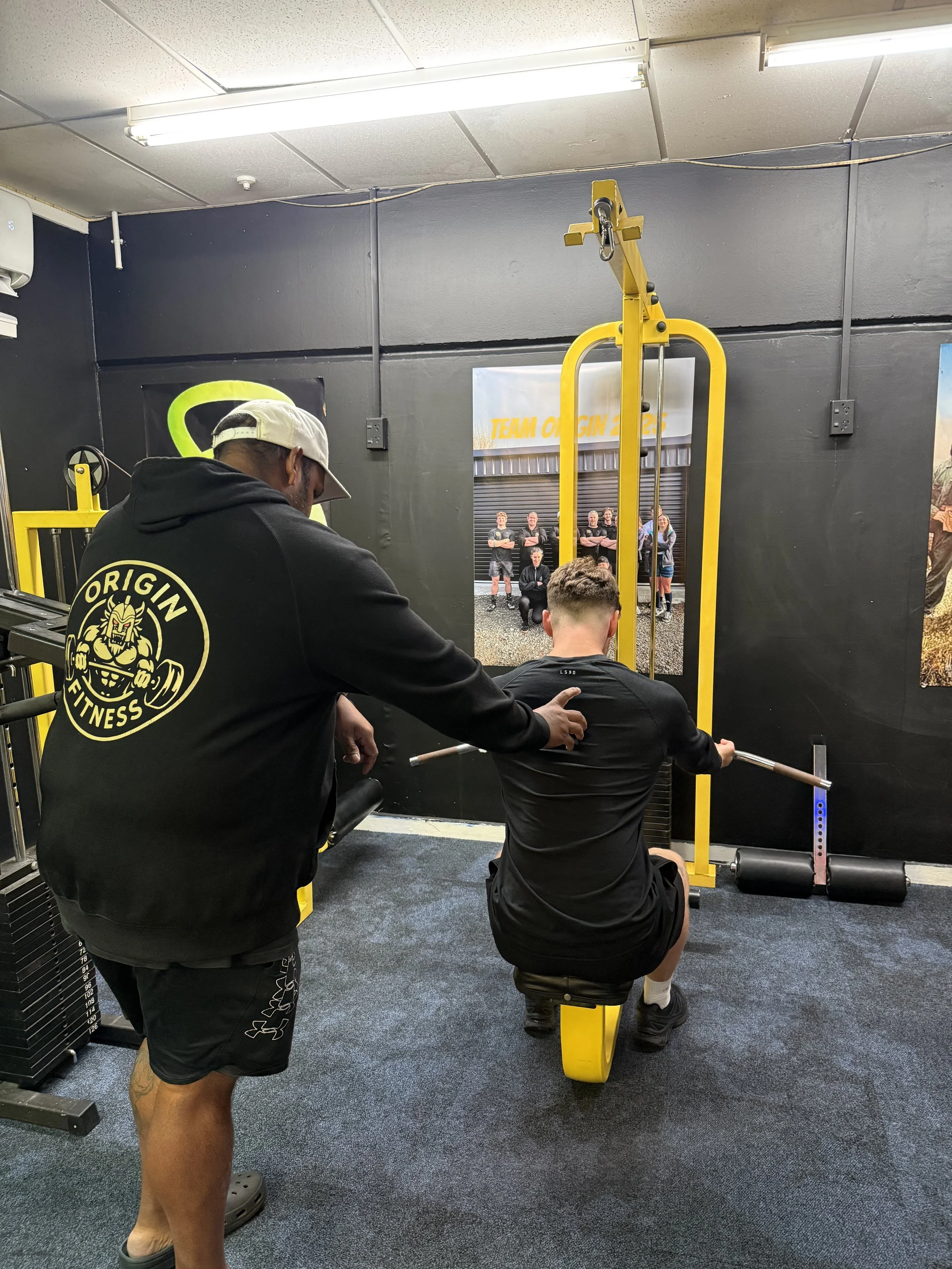 A person sitting on a yellow workout bench lifting a barbell as a trainer stands beside them, giving instructions, inside a gym with black walls and yellow exercise equipment.