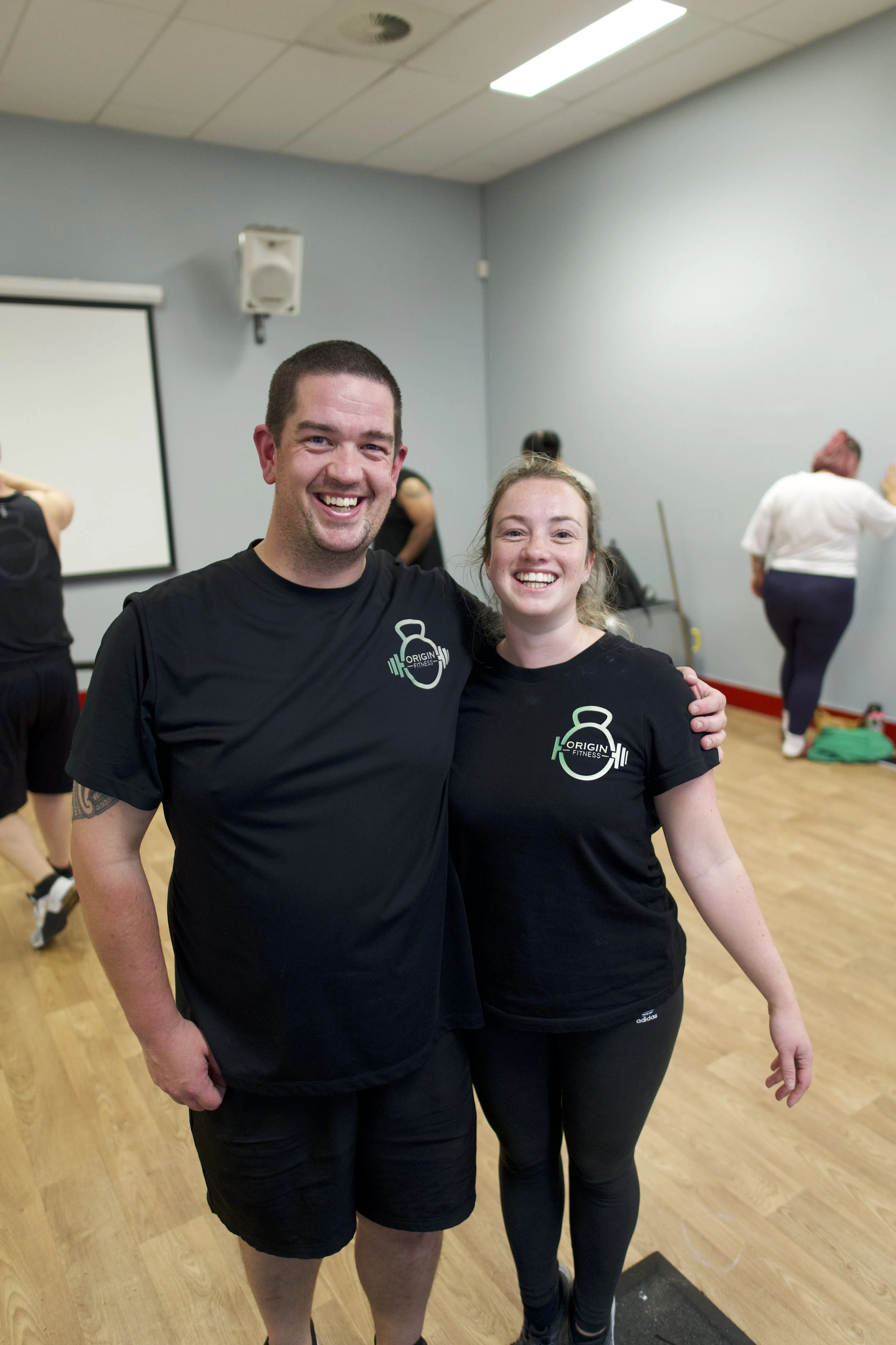 Two smiling individuals wearing black T-shirts with 'Origin Fitness' logos, standing in a room with a group of people exercising in the background.