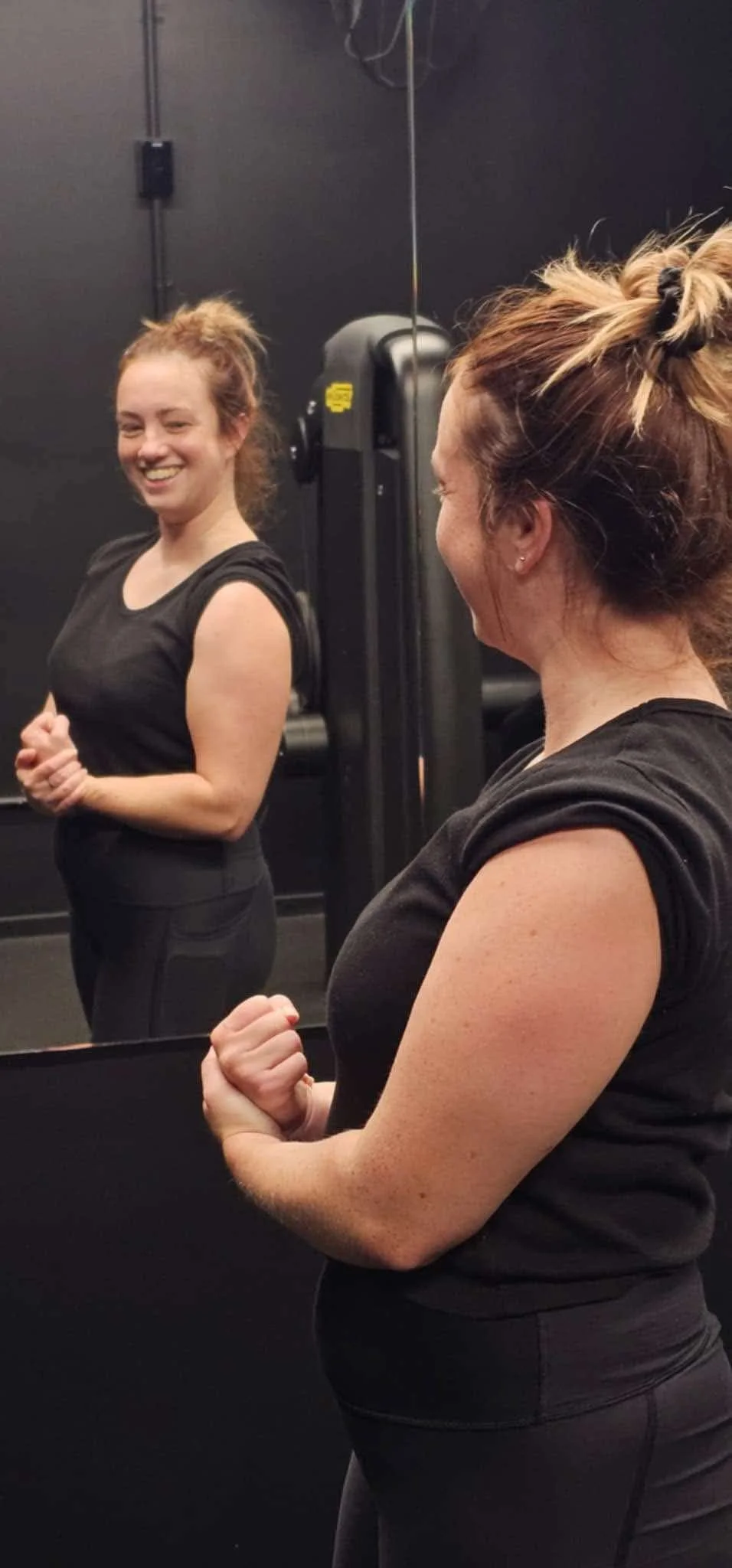 A woman smiling and flexing her bicep while looking at her reflection in a mirror in the gym.
