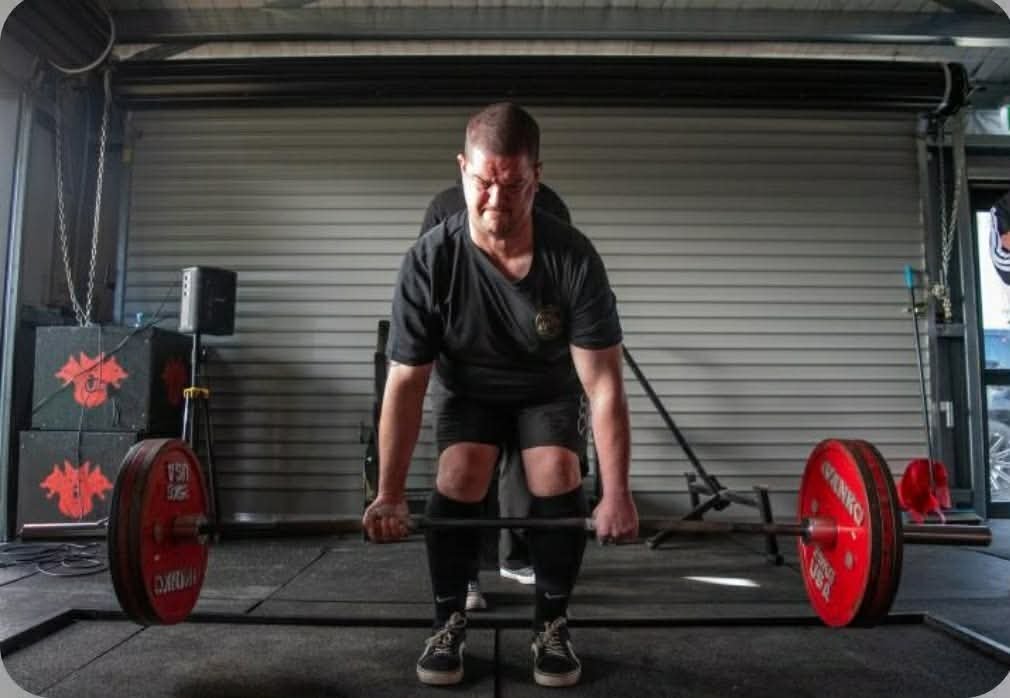 A man in black clothing lifting a barbell with red weights in a gym. The background has a gray roll-up door and workout equipment.