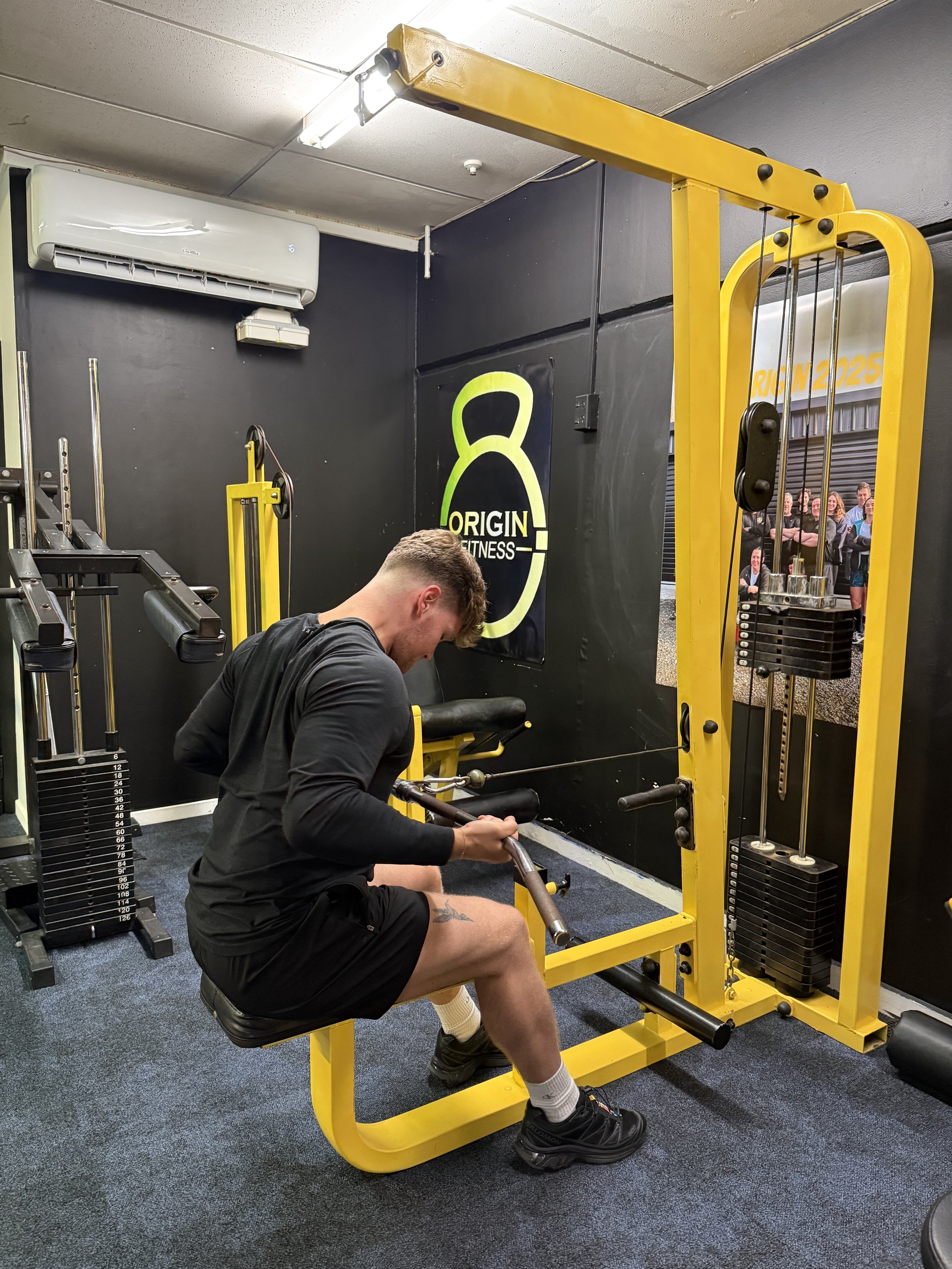 A man in a black athletic shirt and shorts seating on a yellow workout machine, pulling a resistance band with weights attached, inside a gym with black walls and fitness posters.