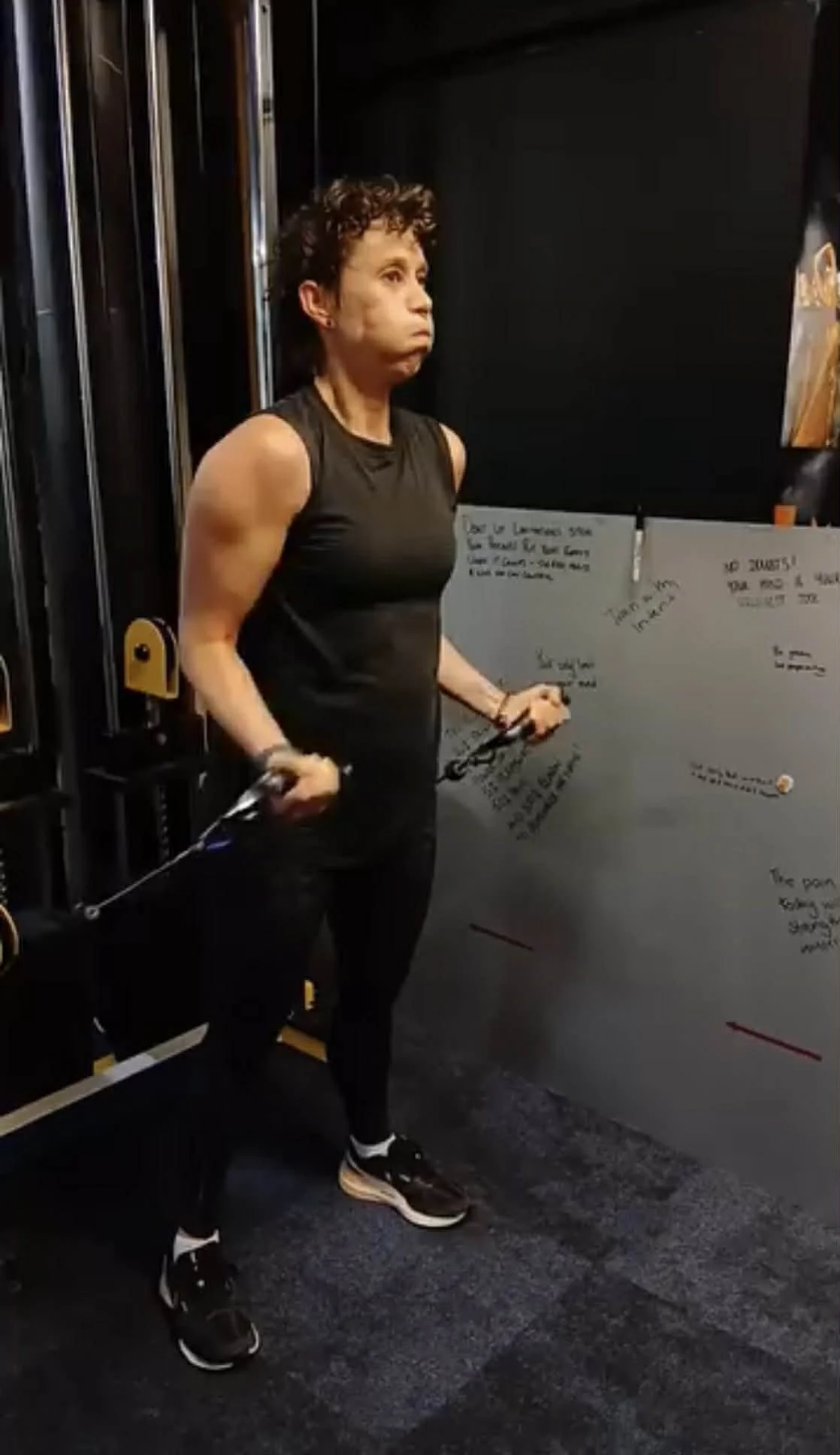 A woman working out with resistance bands in a gym, standing next to a black wall with handwritten notes, wearing a black sleeveless top and black workout pants.