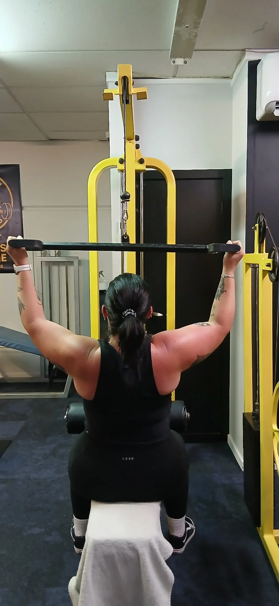 Woman lifting a barbell while seated on a workout bench in a gym.