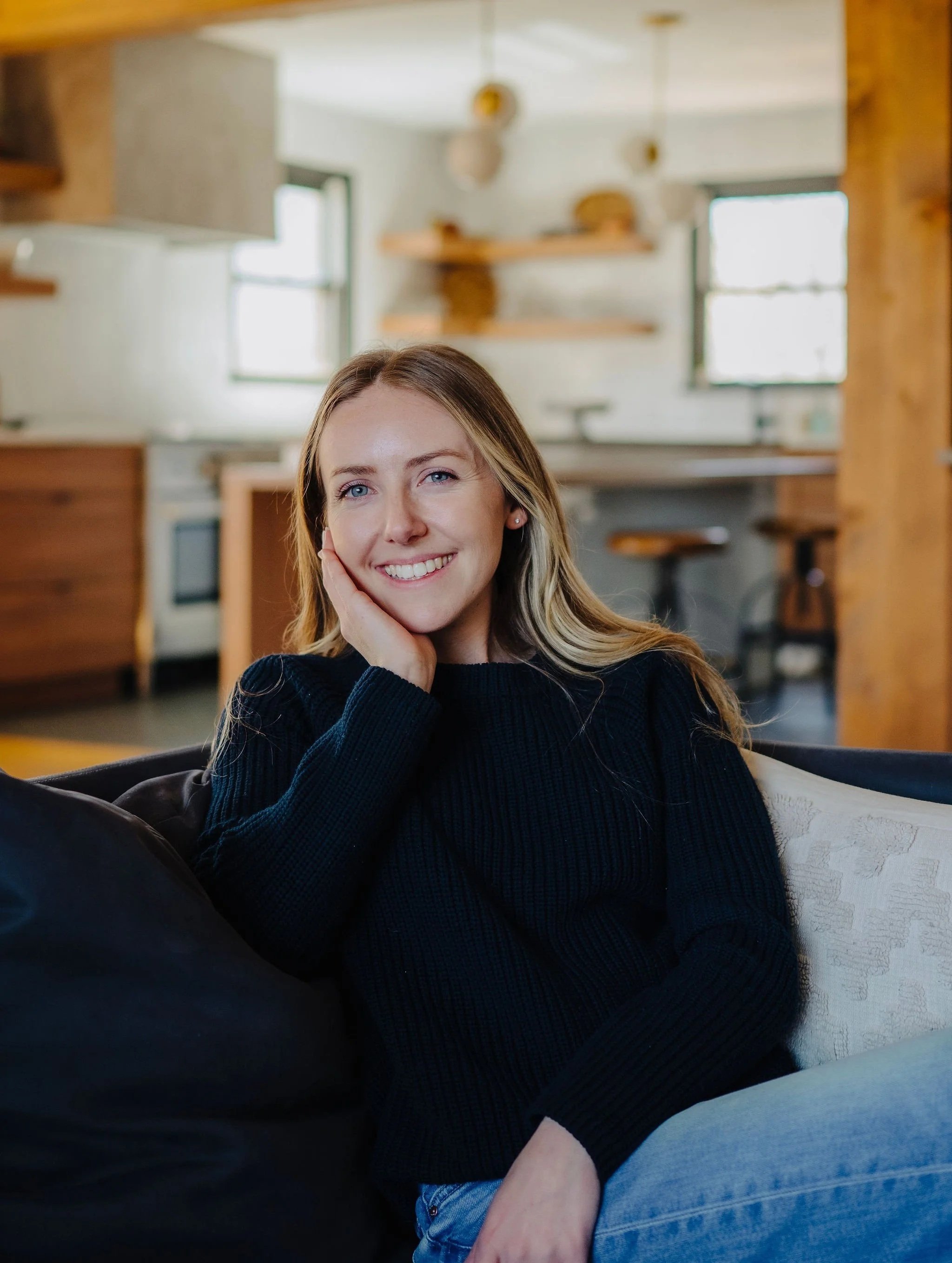 Melanie wearing a black sweater, sitting on a sofa in a cozy living room, smiling with her hand resting on her cheek.