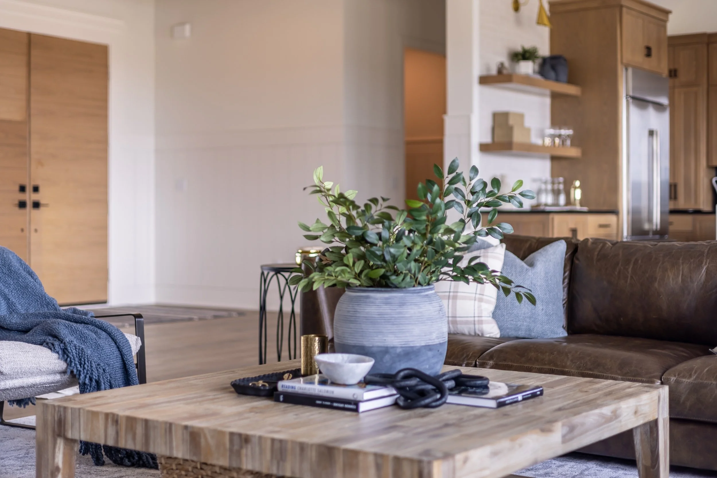 Living room with a wooden coffee table, a potted plant, books, a bowl, and a pair of glasses. A leather sofa with pillows and a throw blanket. In the background, part of the kitchen with wooden cabinets and stainless steel appliances.
