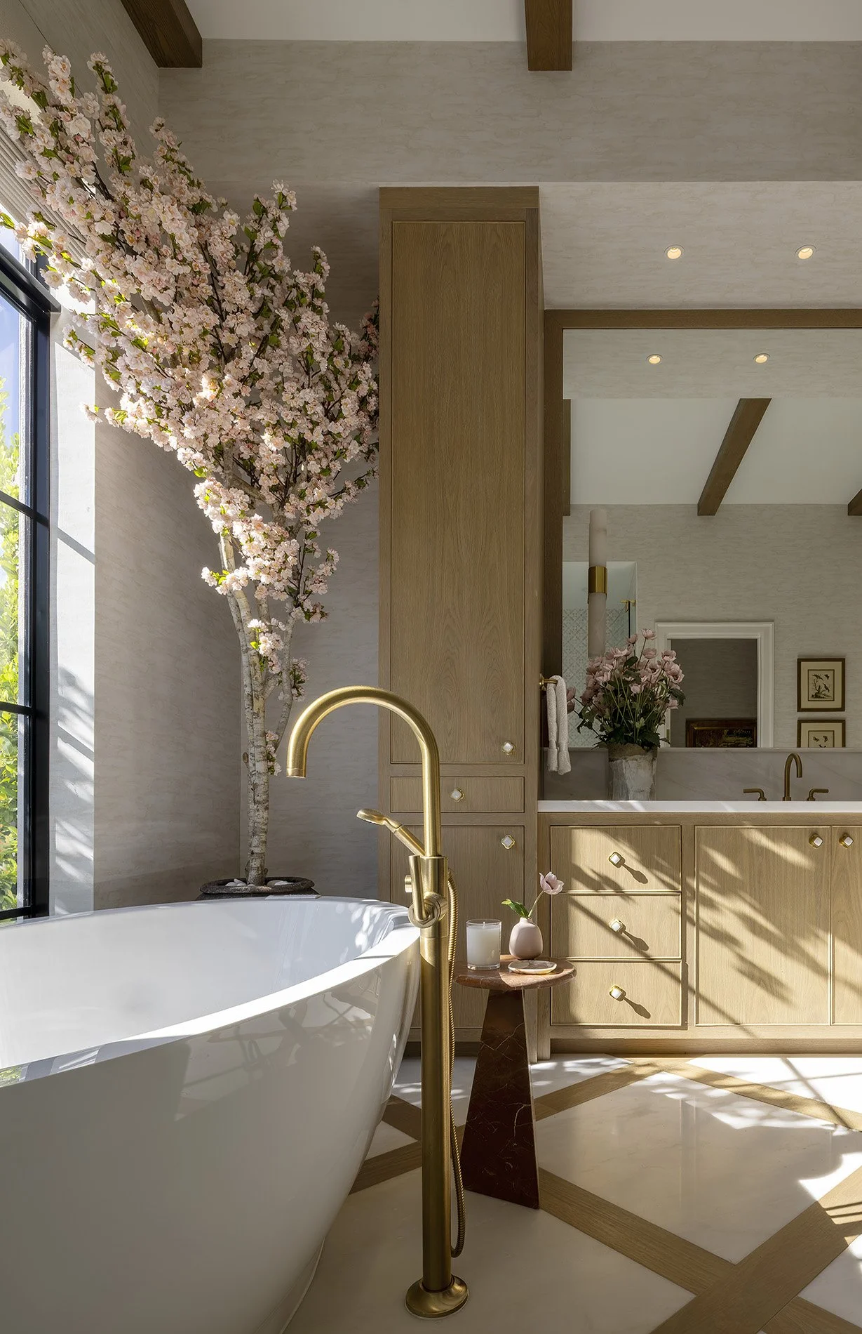 A bathroom interior with a freestanding bathtub, gold-colored faucet, cherry blossom tree, wooden cabinetry, and decorative flowers.