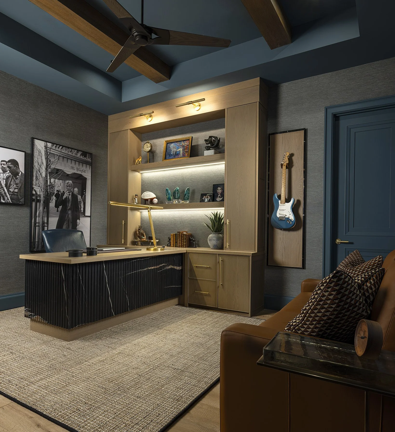 A modern home office with a black and wood desk, gray walls, framed black and white photography, a wooden built-in bookshelf with decorative items, a blue electric guitar mounted on the wall, a brown leather armchair with patterned pillows, and a bei