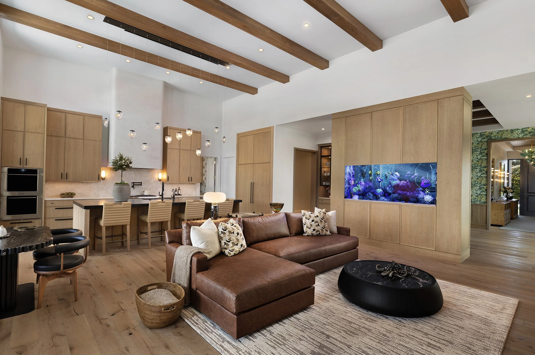 Open-concept living room and kitchen with wooden beams on ceiling, large aquarium built into wall, leather sofa with patterned throw pillows, small black marble coffee table, dining area with chairs, modern pendant lights, and hardwood floors.