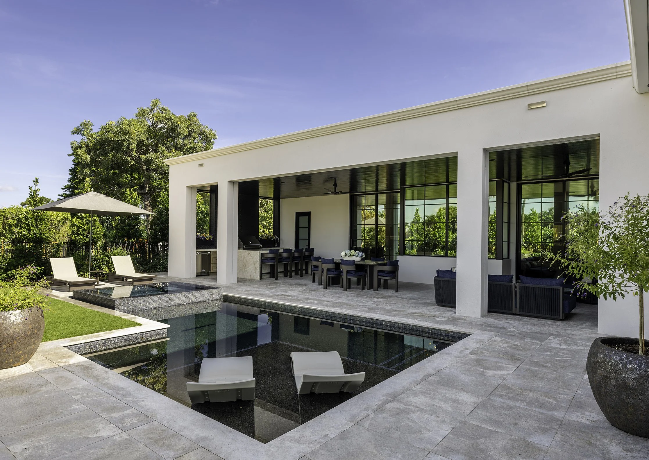 Modern house with large glass windows and a pool in the backyard. There are lounge chairs with white cushions, an umbrella, and lush greenery including trees and potted plants. The sky is clear with some wispy clouds.