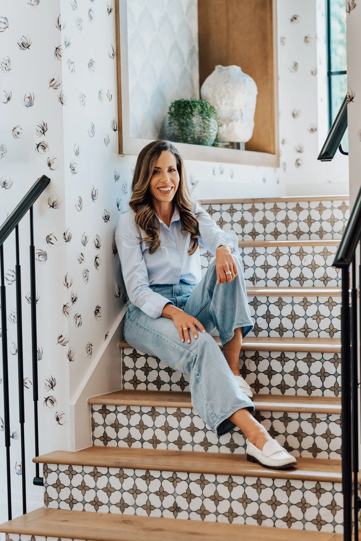 A woman with long wavy brown hair, wearing a light blue shirt and baggy jeans, sitting on a stairway with patterned tiles. She is smiling and looking at the camera. The stairway has wooden steps and a black metal railing. The wall behind her has wallpaper with a spider pattern. A large mirror and a plant are visible in the background.