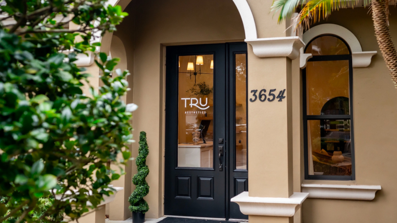 Front entrance door of a building with beige walls, black door with window, house number 3654, and the sign 'TRU Aesthetics' on the door. There are two arched windows on each side of the door, some plants in front, including a potted spiral topiary and a palm tree.