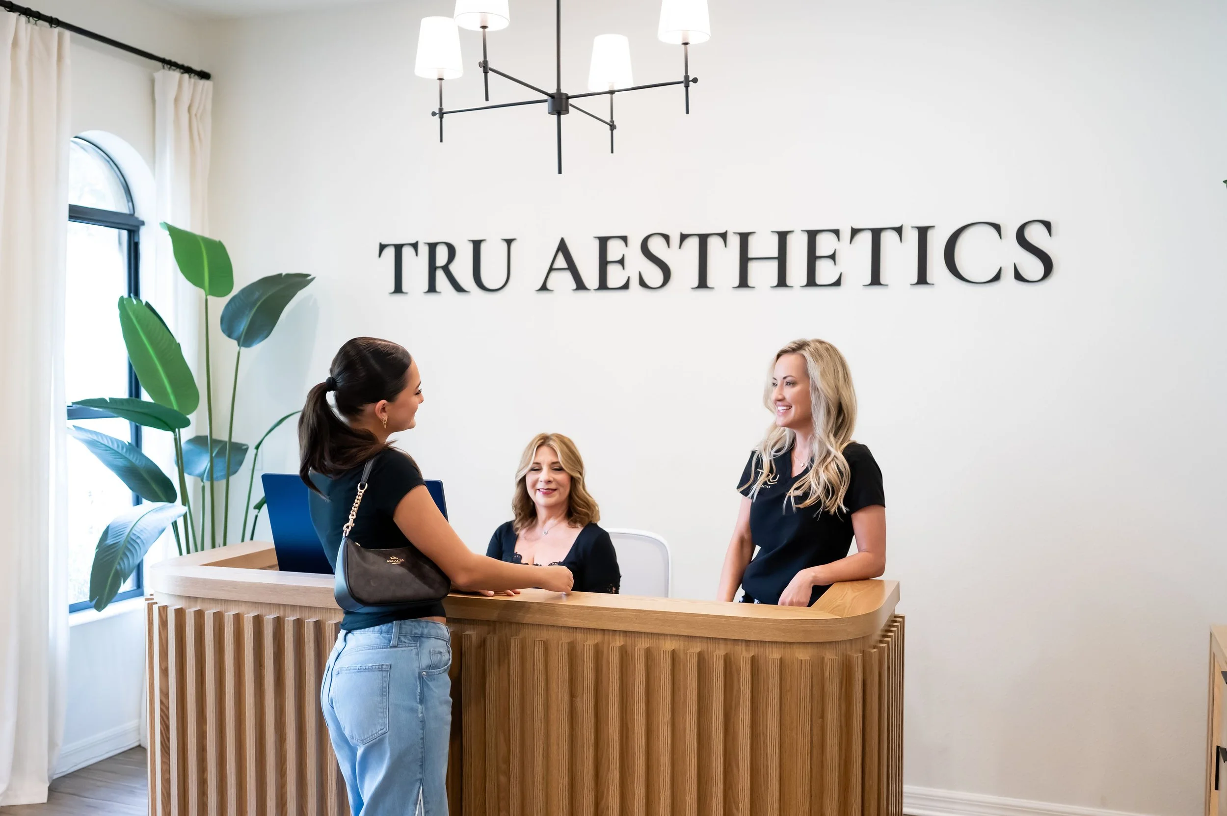 Reception area at Tru Aesthetics with three women interacting, a woman behind the counter, a large green plant, and a white wall with the business name in black letters, modern lighting.