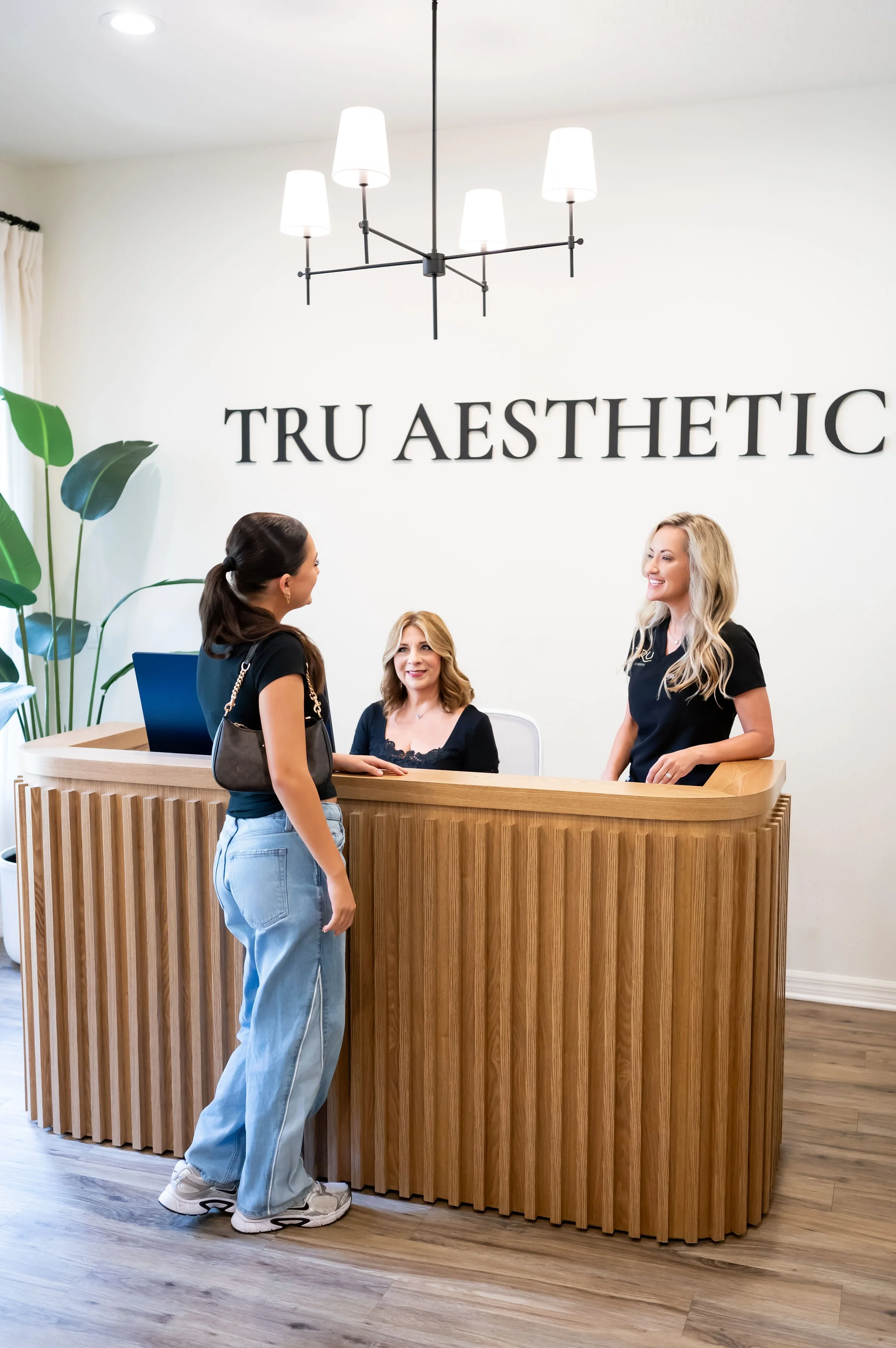 Three women at a reception desk with a sign that reads 'TRU AESTHETIC' and a modern chandelier overhead. One woman is standing and two women are sitting behind the desk, all smiling and engaging in conversation.
