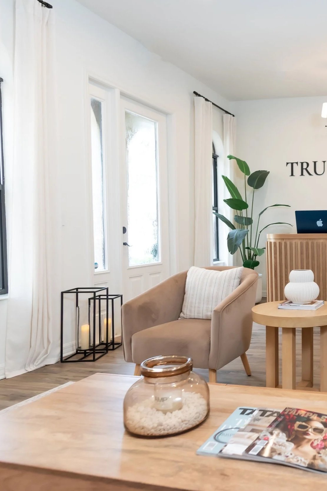 Cozy living room with beige armchair, wooden coffee table with a glass jar filled with decorative white stones, magazines, white walls, large windows with white curtains, green plant, black candle lanterns with candles, and partially visible laptop on a wooden counter.