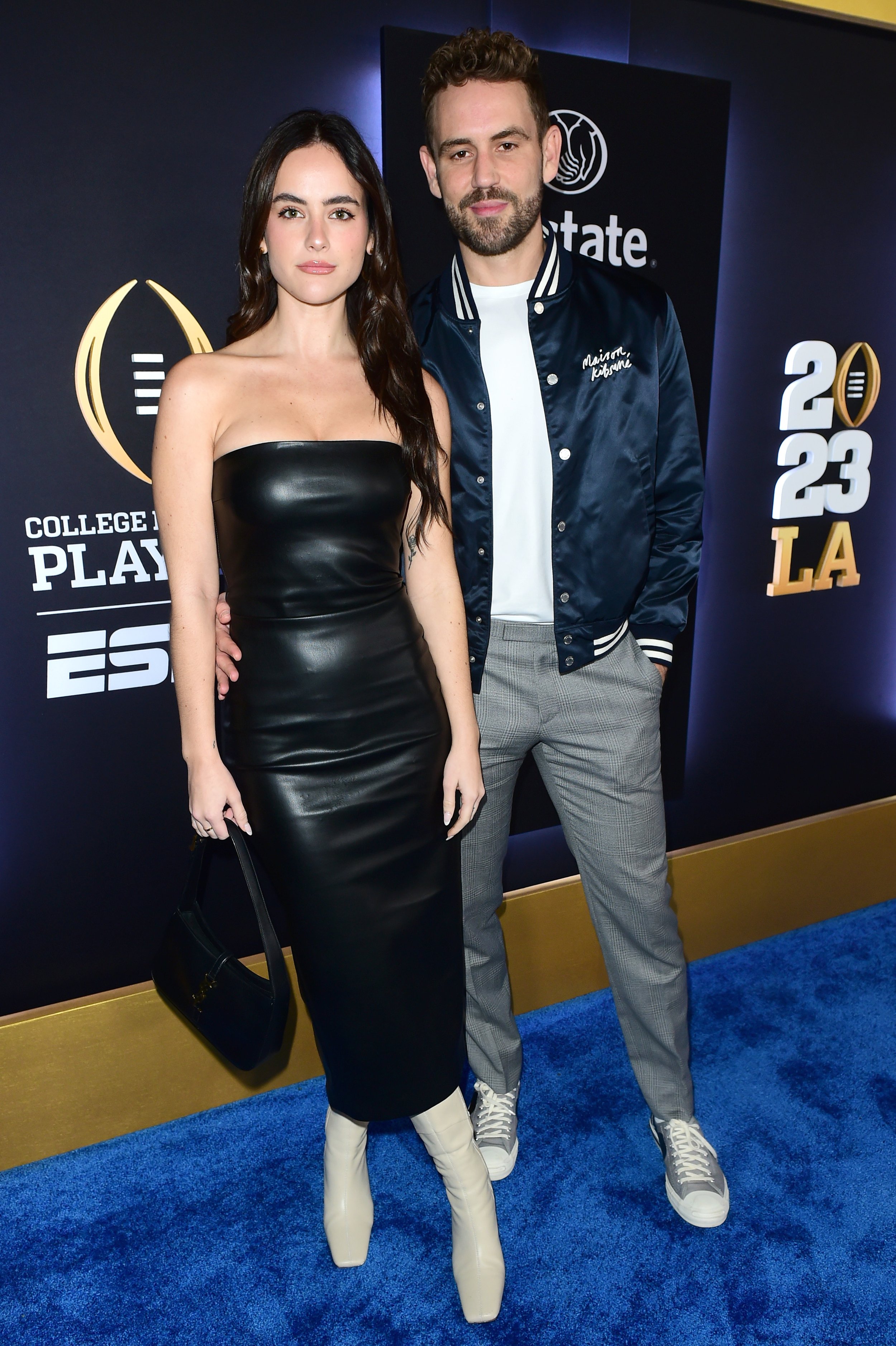 A man and a woman posing together on a blue carpet at the 2023 College GameDay LA event. The woman has long dark hair and is wearing a strapless black leather dress, white high heel boots, and is holding a black handbag. The man has short curly hair and a beard, and is wearing a navy blue satin bomber jacket with white trim, a white T-shirt, gray plaid pants, and gray sneakers.