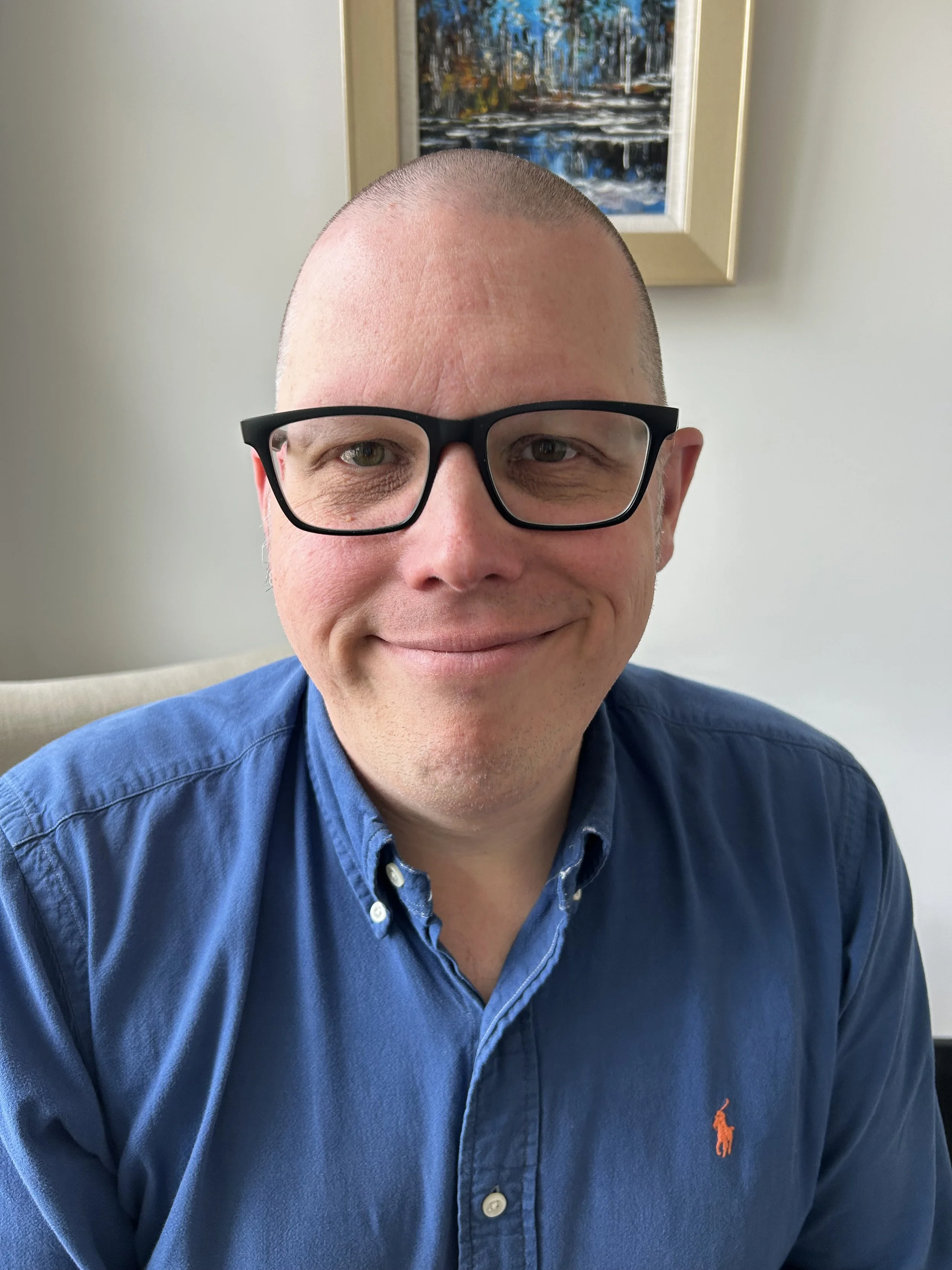 A man wearing black glasses and a blue collared shirt smiling in front of a painting and a white wall.