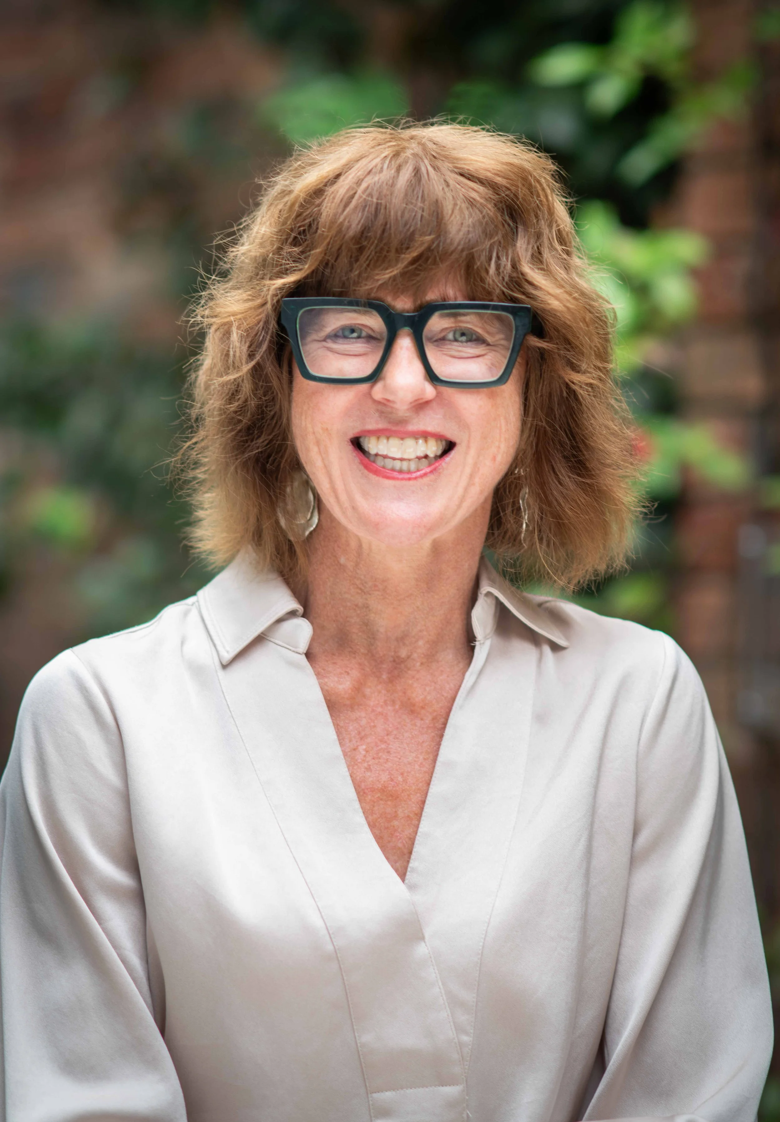 Portrait of Jill Skromanis smiling, wearing black glasses and a light blouse, standing outdoors with greenery in the background.