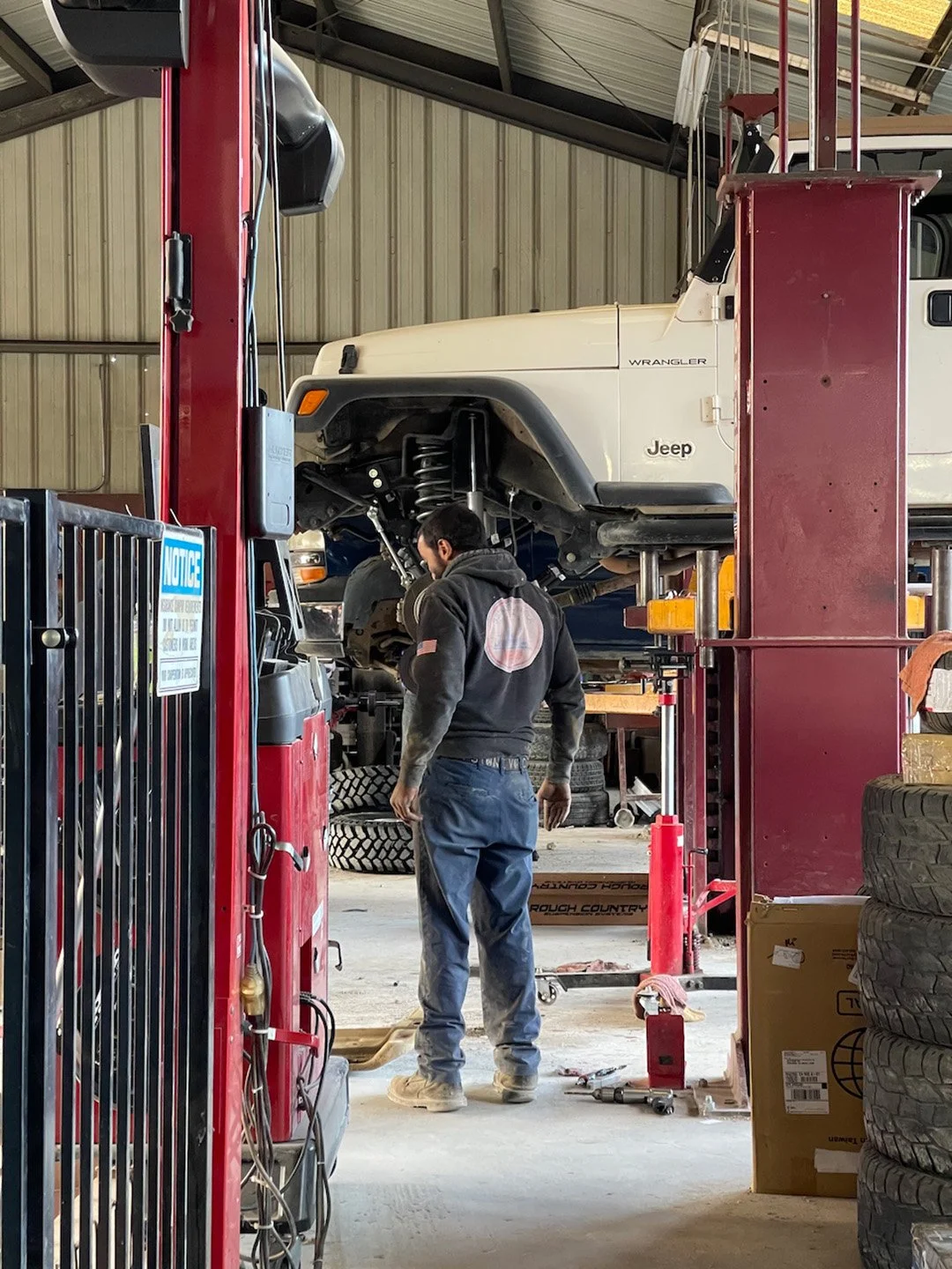 Jeep: “I’ve been through a lot.”
Us: “Good thing you’re here.”

A technician stands beside a lifted Jeep Wrangler inside the shop, with the vehicle raised on the lift and tools nearby, showing a quiet moment of focused work during a regular day in th