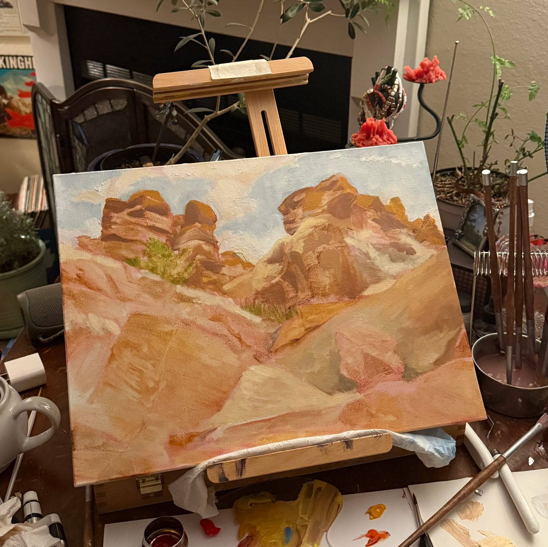An artist's easel holds a partially completed landscape painting of mountains and clouds. Surrounding the easel are various art supplies, plants, and a candle on a cluttered table.
