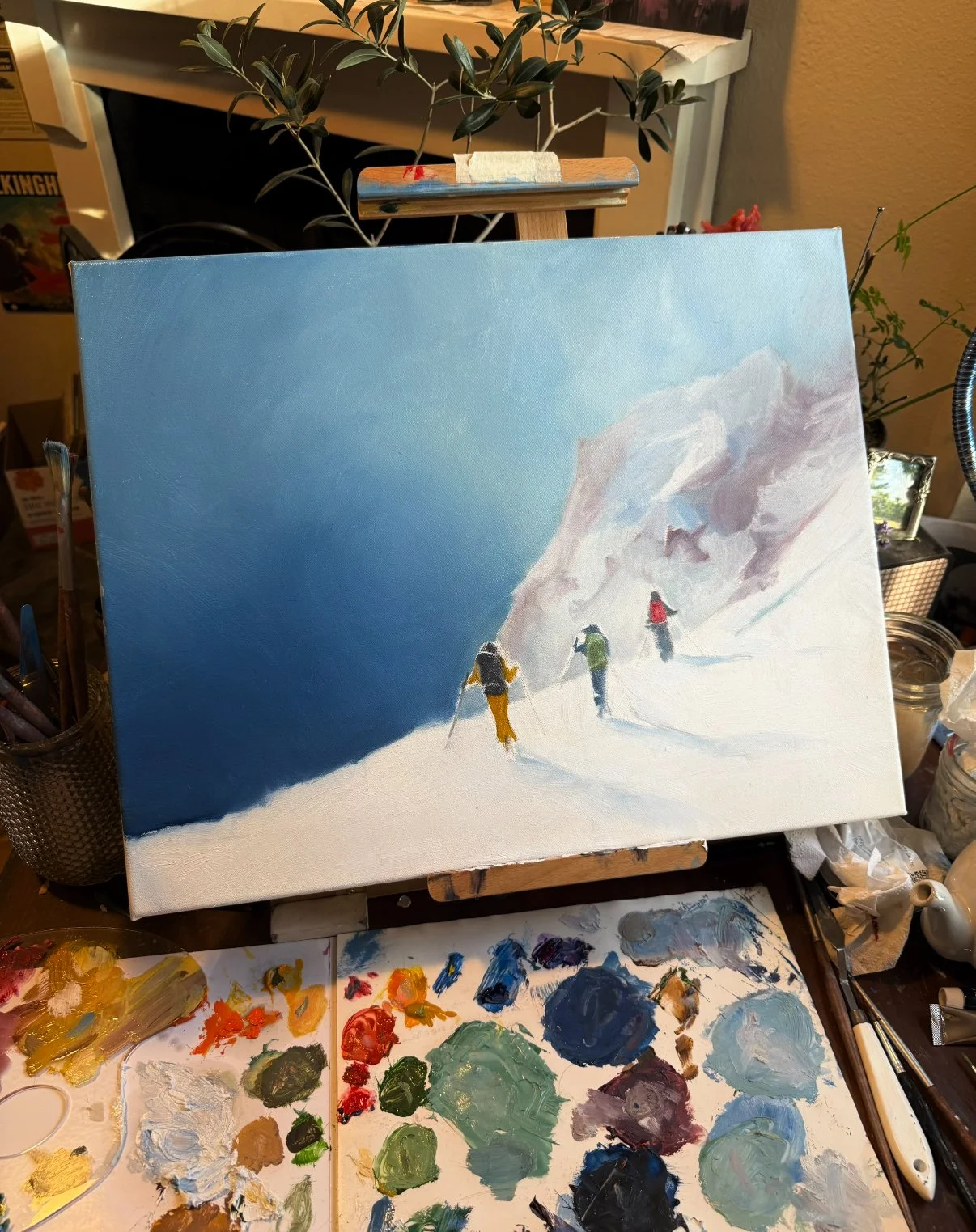 An unfinished landscape painting of a snowy mountain scene with three hikers in colorful jackets, on an artist's workspace with paint palettes and brushes.