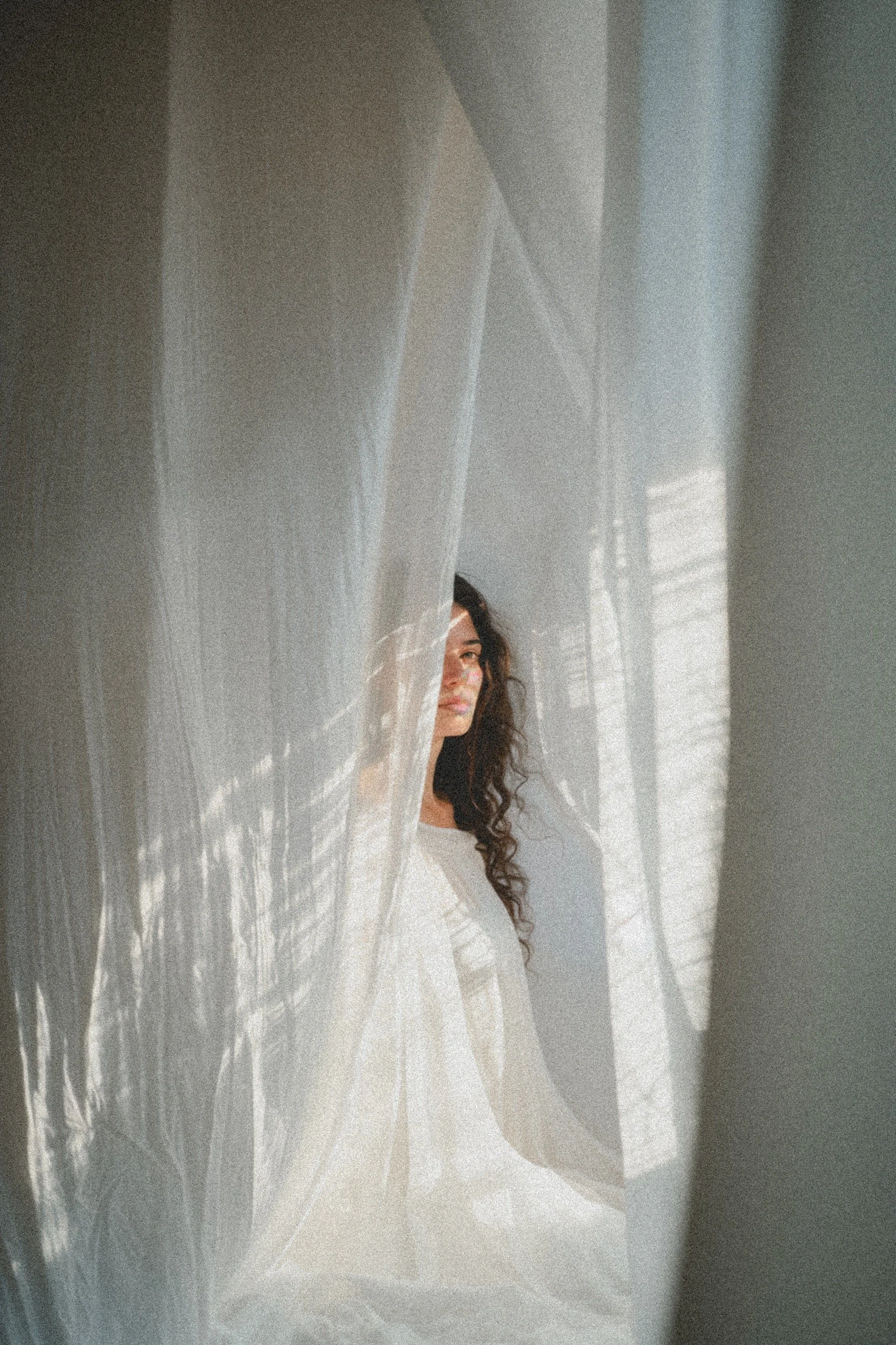 A woman with curly hair peeking out from behind a semi-transparent curtain with sunlight streaming through.