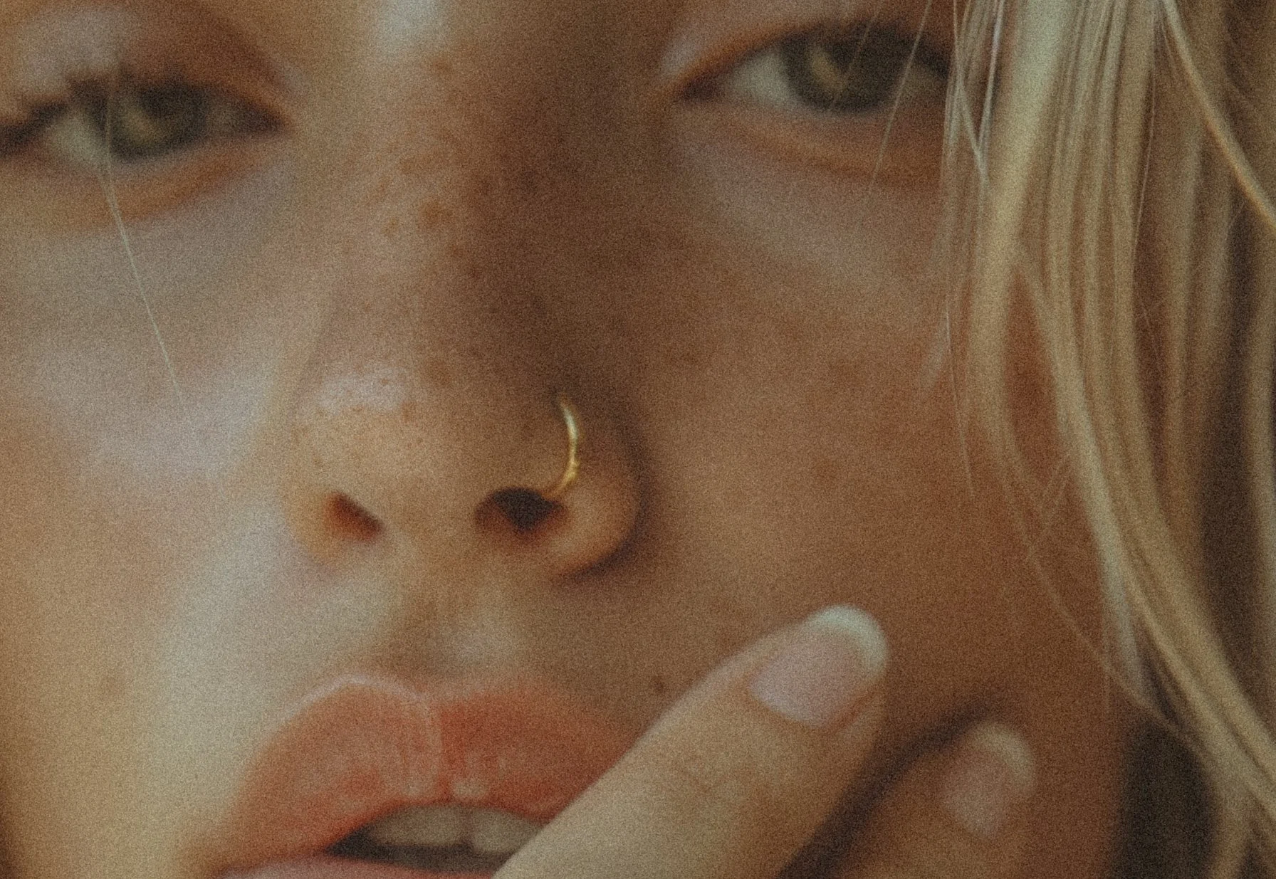 Close-up of a person's face showing their eye, nose, and lips, with a finger touching their chin. The person has a gold nose ring and long blond hair, with some background tiles visible.