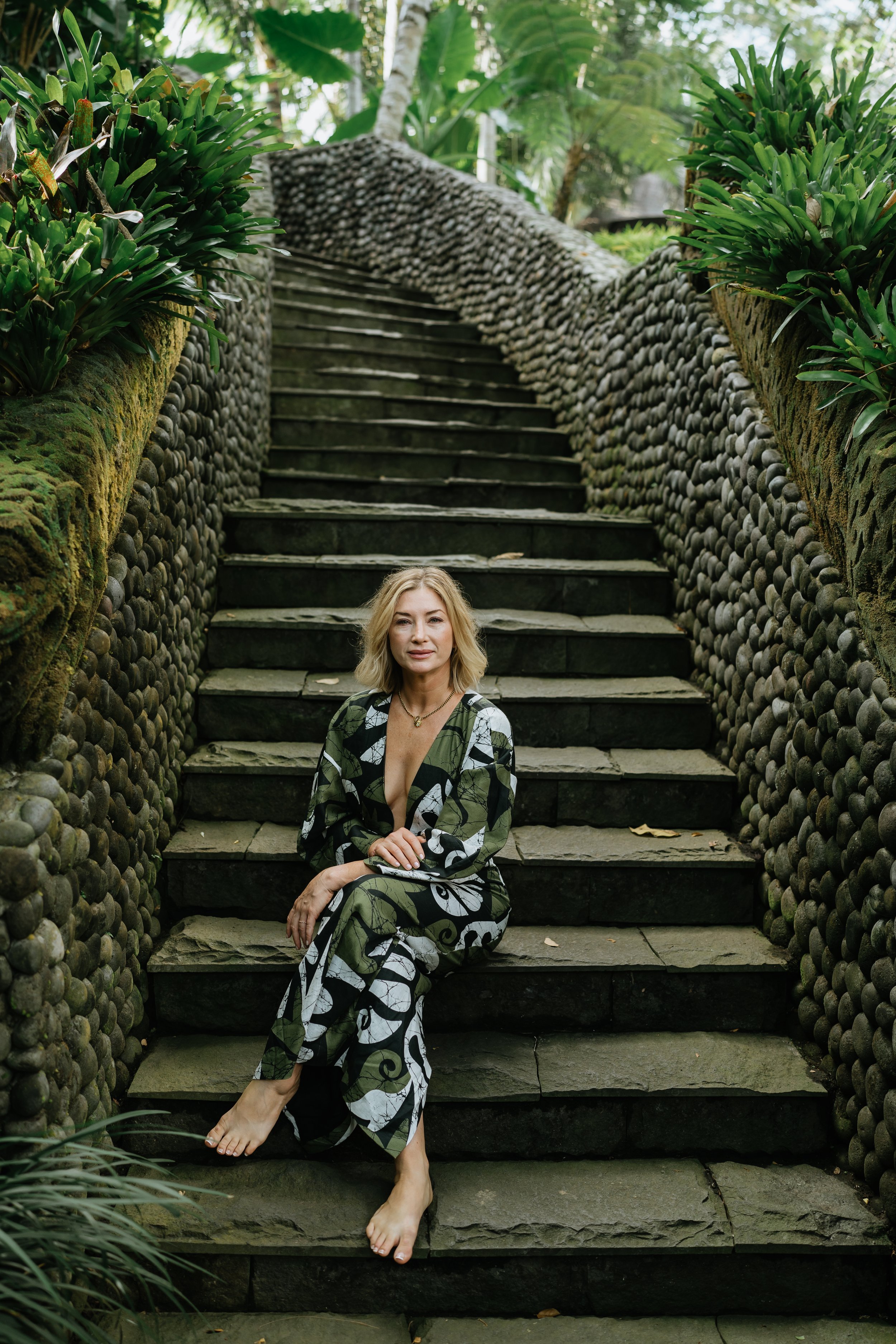 A woman with blonde hair sitting on stone stairs in a lush, green outdoor setting, wearing a patterned dress and barefoot.