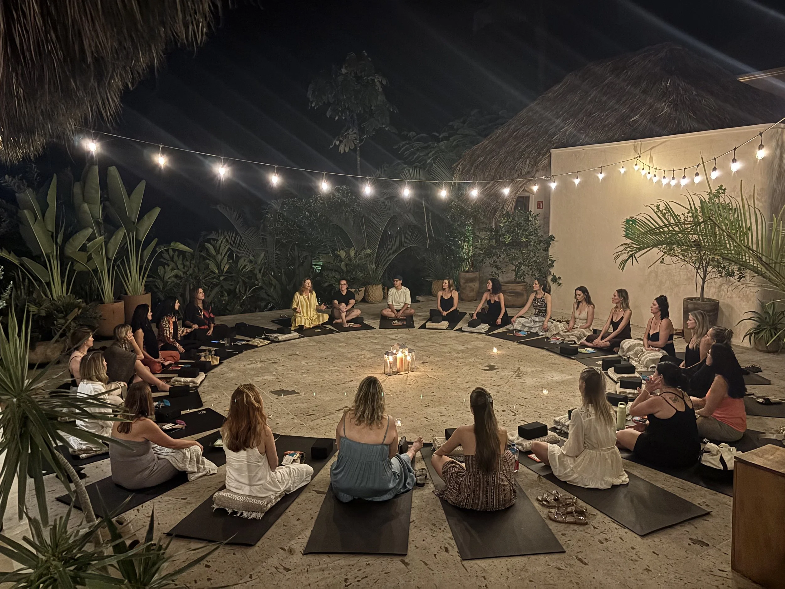 A group of people sitting cross-legged on yoga mats outdoors at night, participating in a meditation session surrounded by lush greenery and tropical plants, illuminated by string lights overhead.