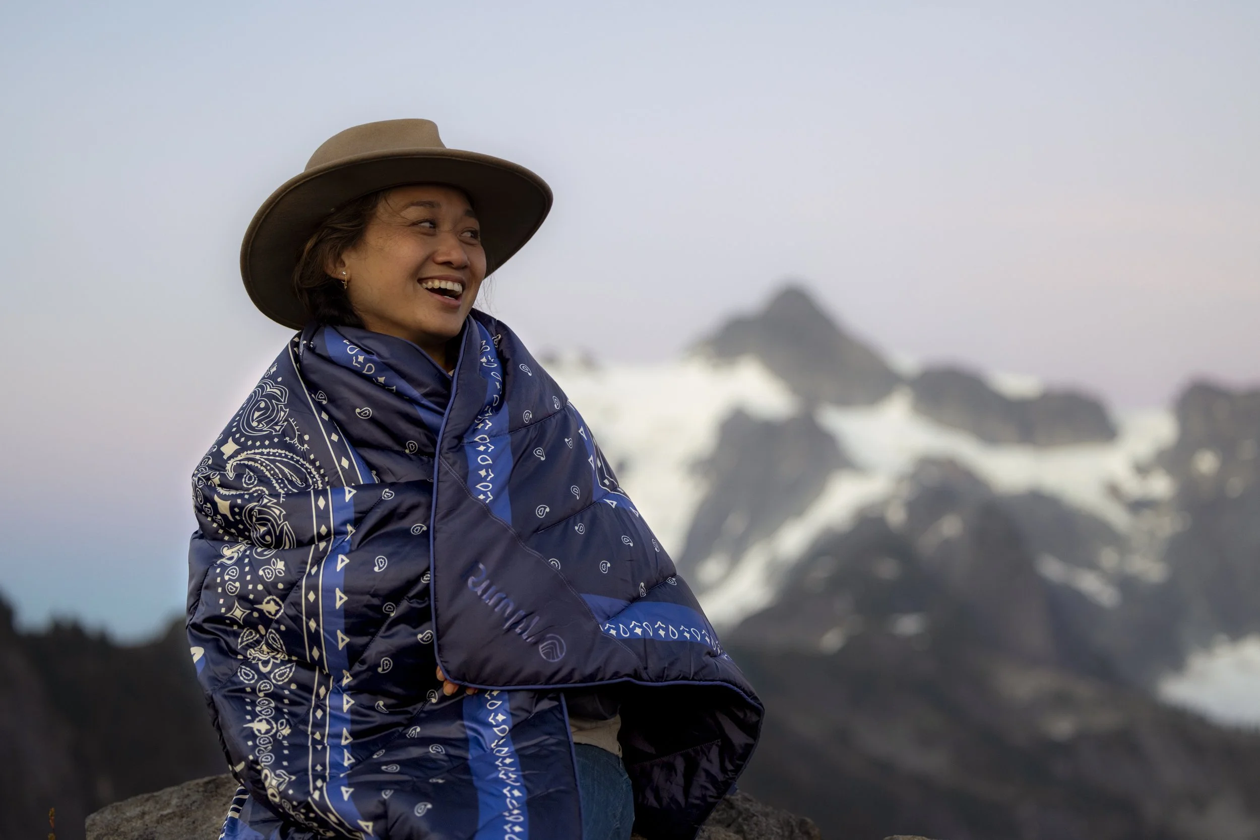 A woman in a blue patterned jacket and a wide-brimmed hat smiling outdoors with snow-capped mountains in the background.