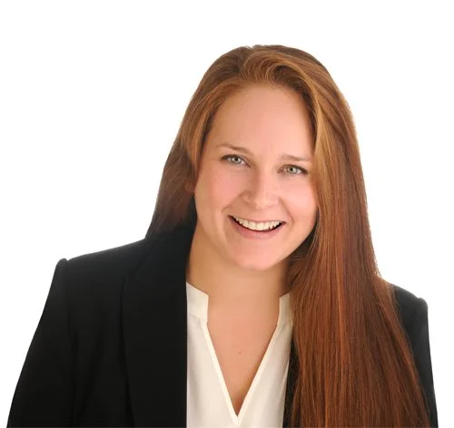 A smiling woman with long red hair wearing a black blazer and white blouse against a white background.