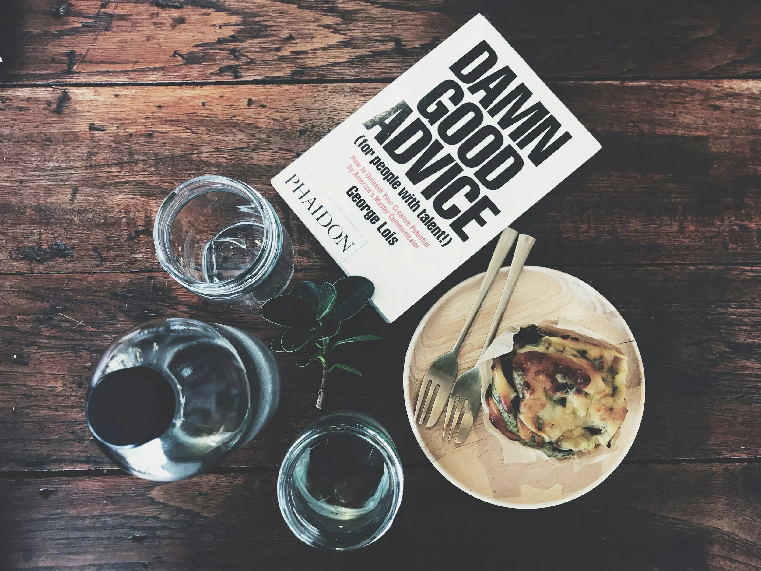 A wooden table with a book titled 'Damn Good Advice' by George Lois, two glasses of water, a small leaf, a small plate with a slice of pizza, and two forks.