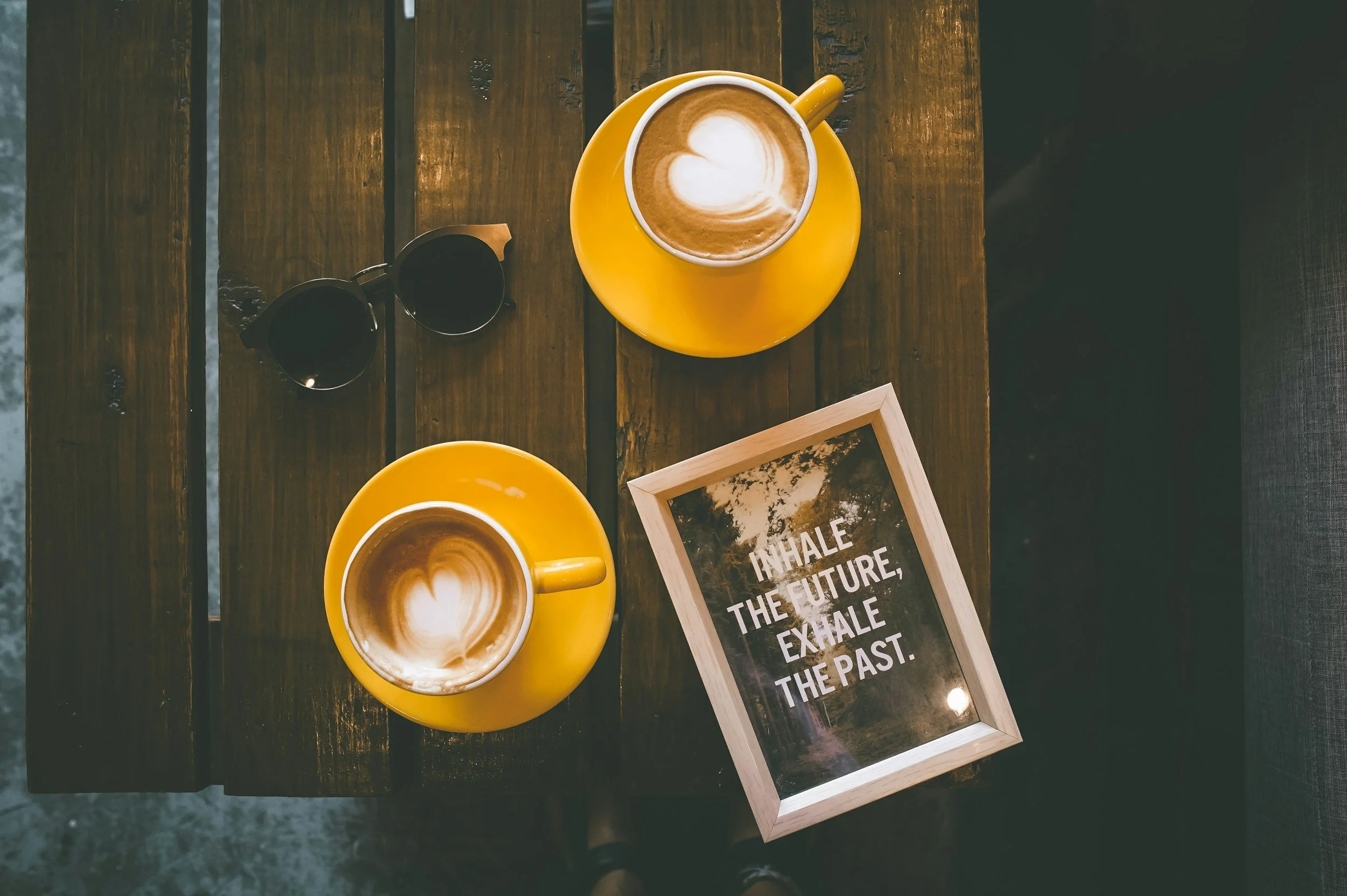 Two yellow plates with coffee cups and latte art, a pair of black sunglasses, and a framed quote on a wooden table. The quote says, 'Inhale the future, exhale the past.'