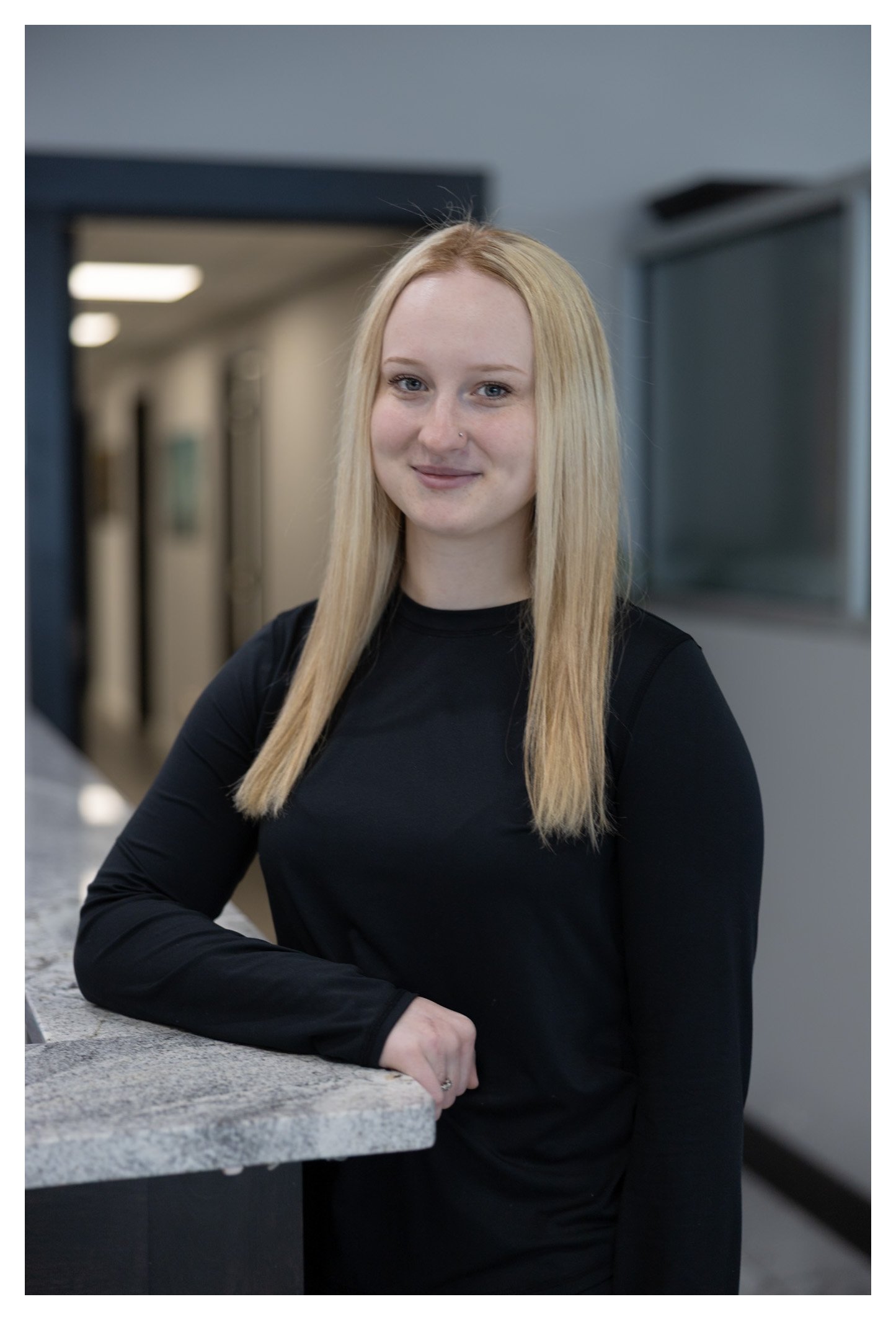 A young woman with blonde hair and a nose piercing, wearing a black long-sleeve shirt, standing at a countertop in an indoor setting, smiling at the camera.