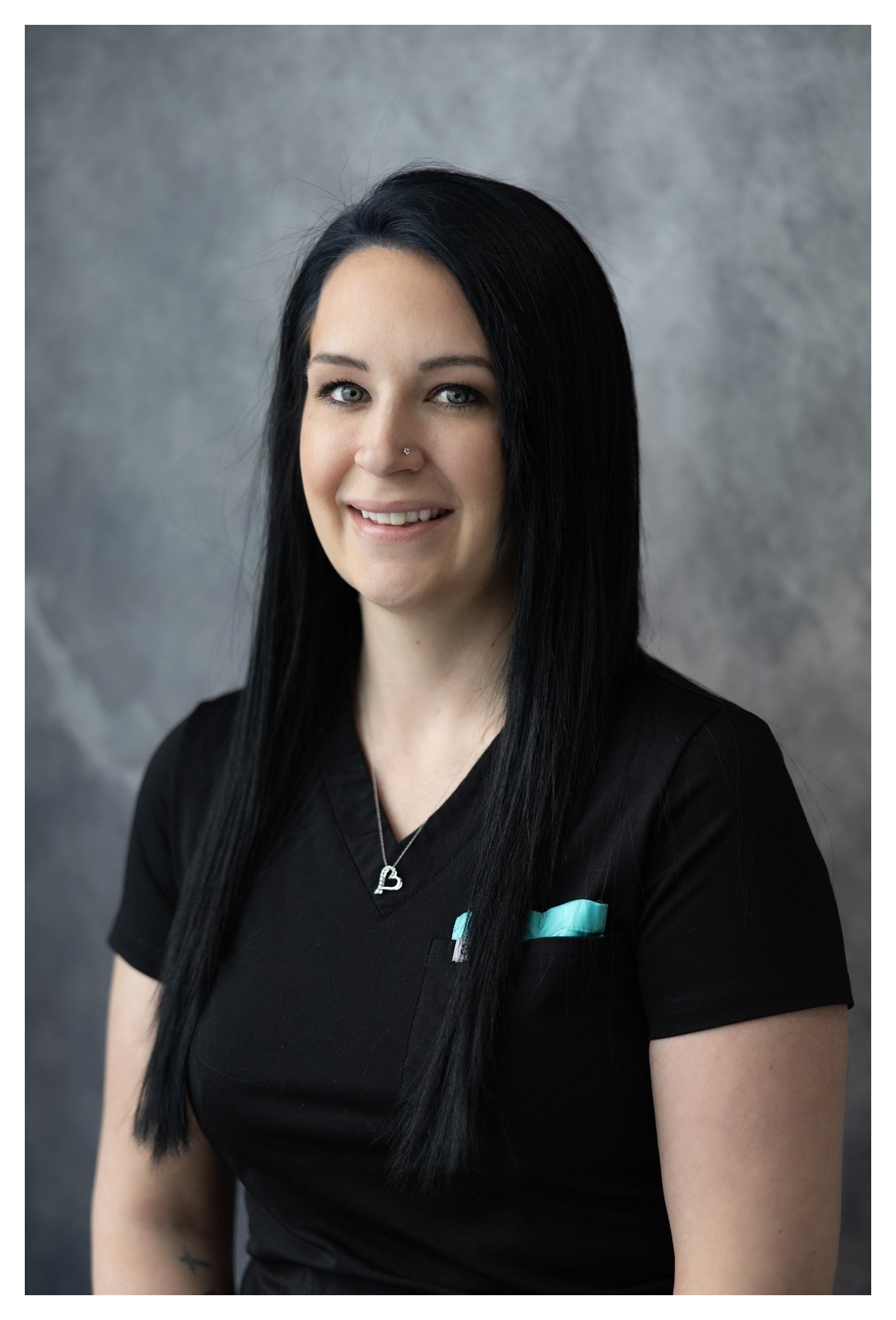 A woman with long black hair, fair skin, and blue eyes, smiling slightly, wearing a black top with a light blue detail and a silver necklace with a heart-shaped pendant, standing against a gray textured background.