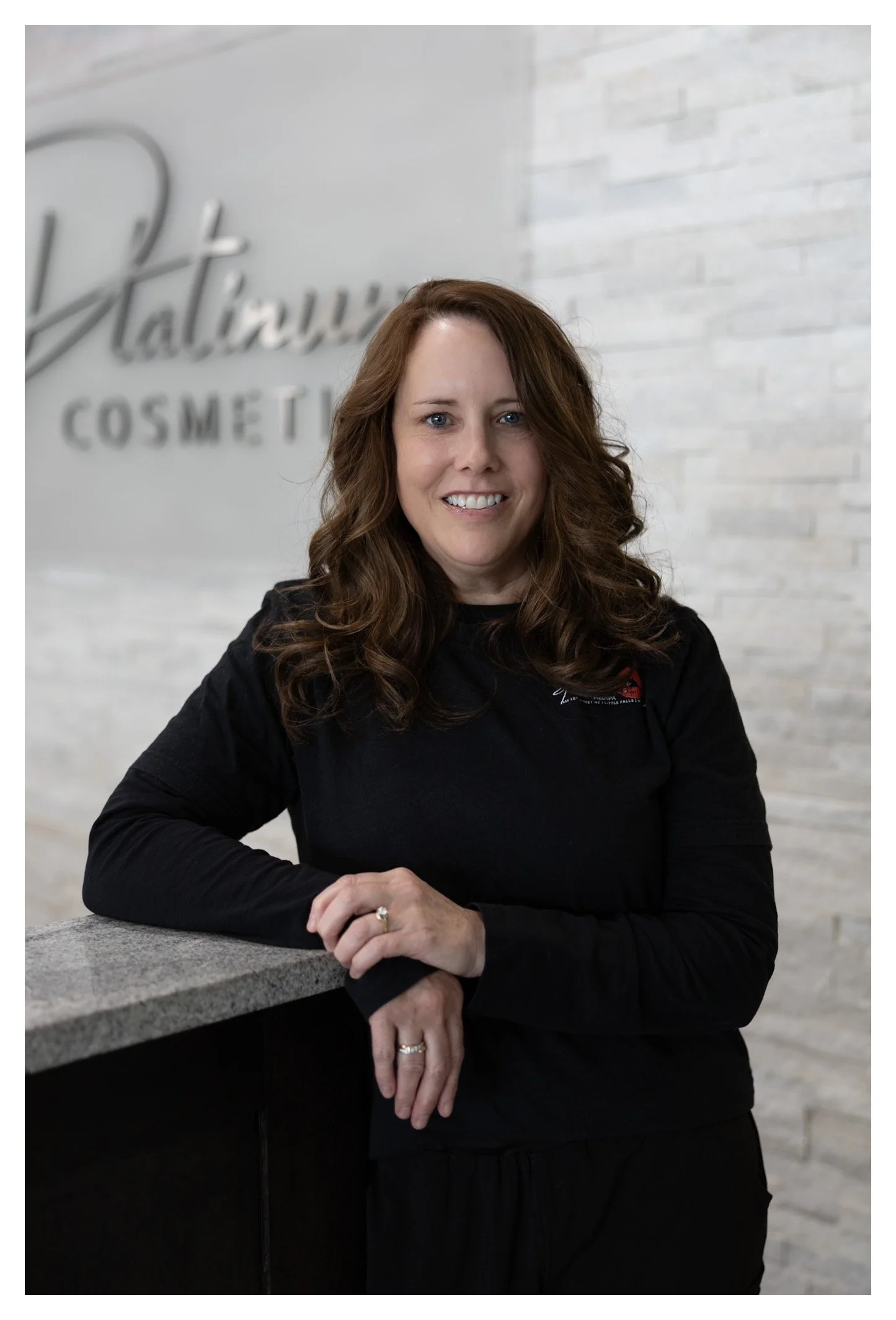 Woman with brown curly hair smiling in front of a white wall with a logo that reads "Patricia Cosmetics."
