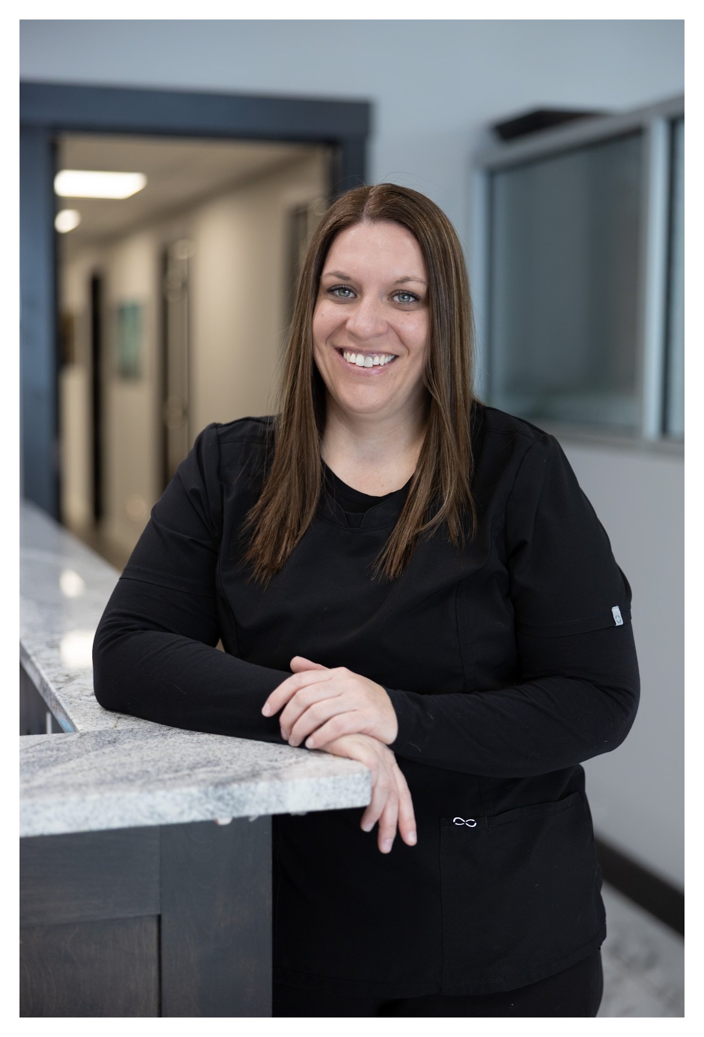 A smiling woman with long brown hair, dressed in black scrubs, leaning on a granite countertop in a professional office environment.