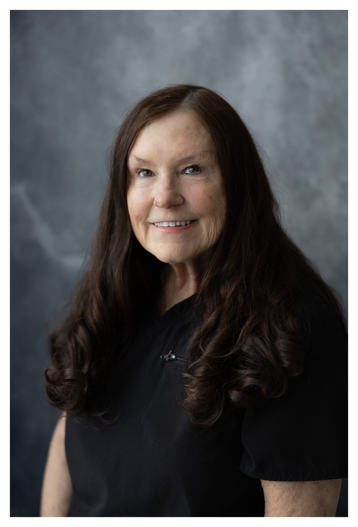Portrait of a woman with long, wavy brown hair, wearing a black top, smiling at the camera against a gray textured background.