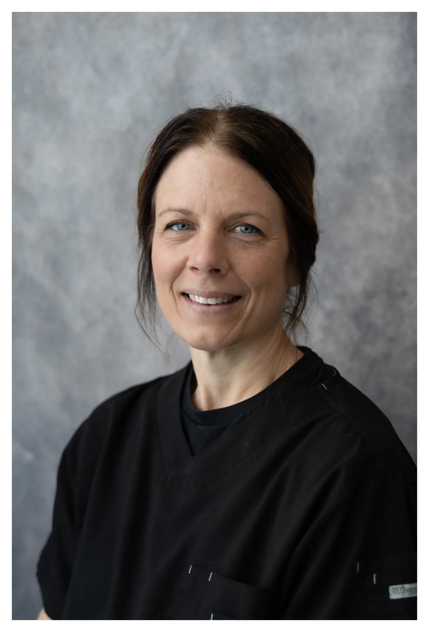 A woman with short dark hair, blue eyes, and fair skin wearing a black medical scrub top, smiling at the camera, against a gray textured background.