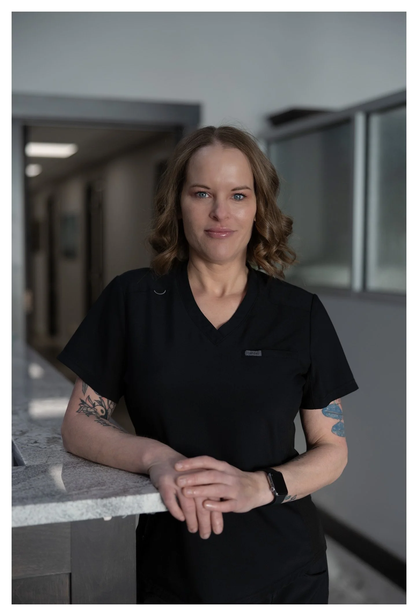 A woman in black scrubs with tattoos on her arms, standing indoors by a countertop, looking at the camera with a slight smile.