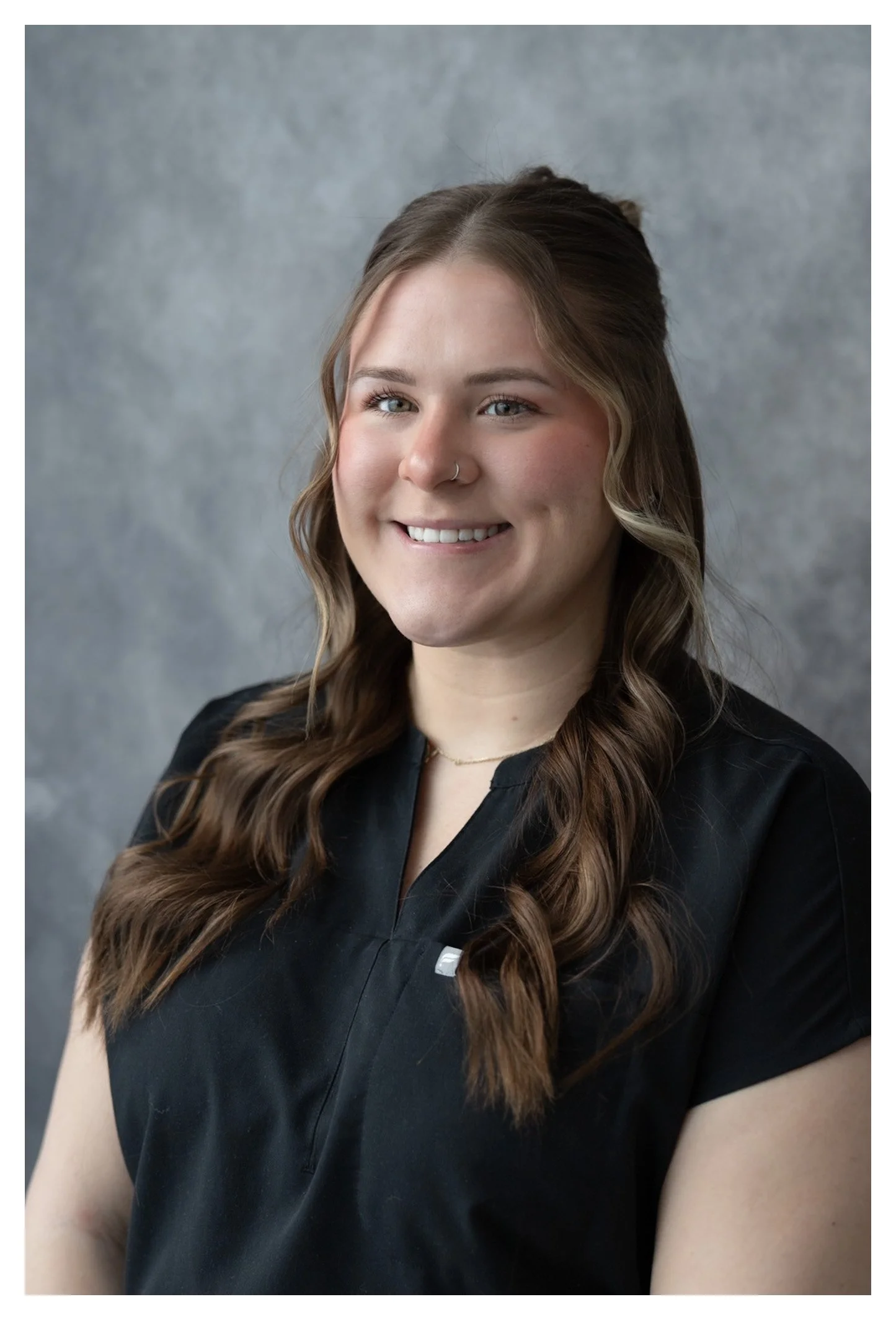 A young woman with long, wavy brown hair, smiling, wearing a black top, and standing against a gray background.