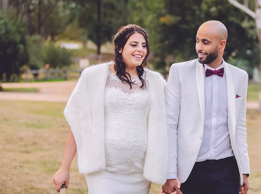 A bride and groom holding hands and walking outdoors, smiling at each other. The bride is wearing a white lace wedding dress and a white fur shawl, and the groom is dressed in a white tuxedo with a maroon bowtie.