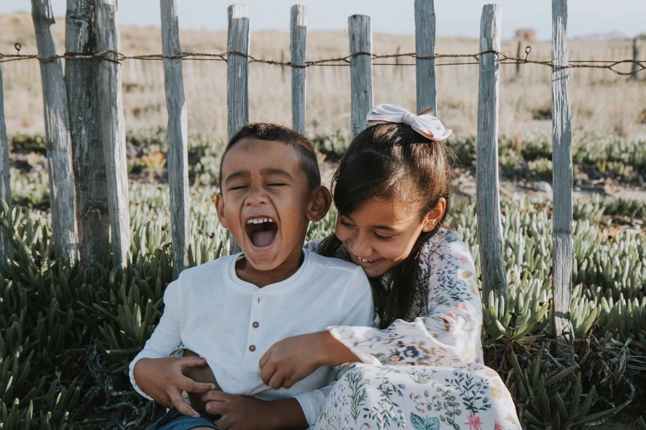 Two children sitting outdoors behind a wooden fence, laughing and enjoying each other's company, with plants in the background.